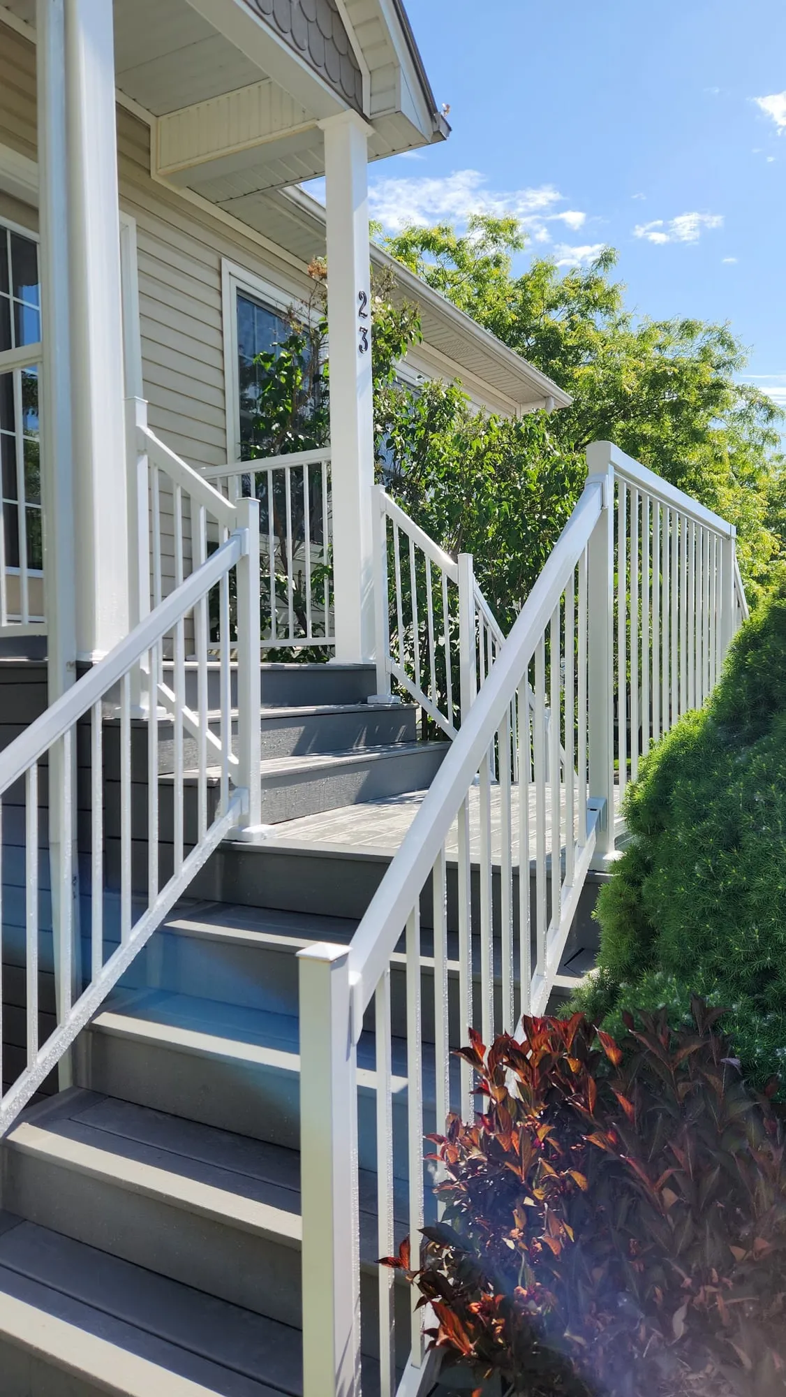 A finished front stairway with grey treads and white railings climbing to a covered porch beside green shrubs.