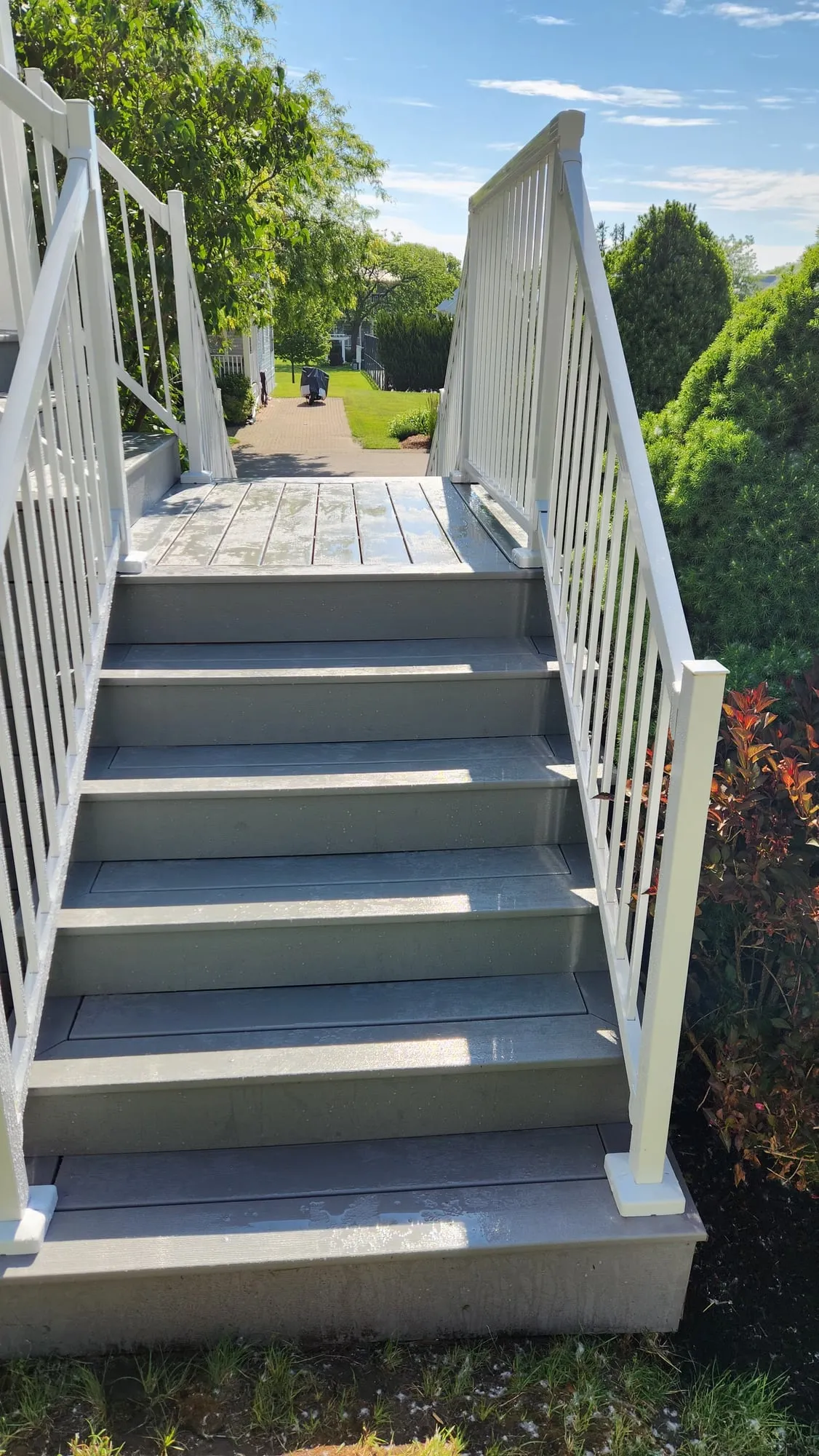 A grey staircase with white railings rising to a landing, with a paved path and green lawn beyond.