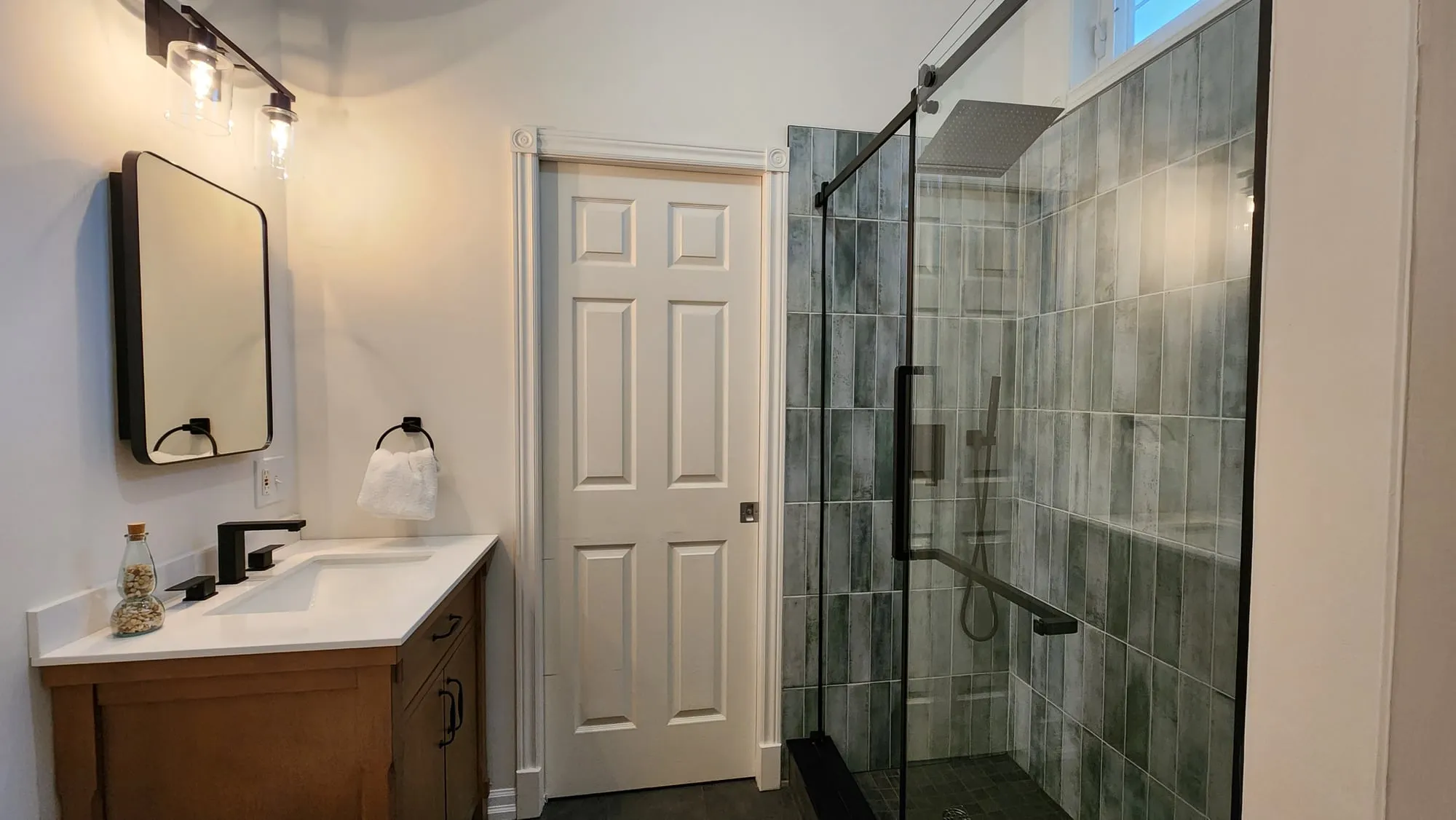 A finished bathroom with a wood-toned vanity, black-framed mirror and a glass shower lined in grey-green tile.