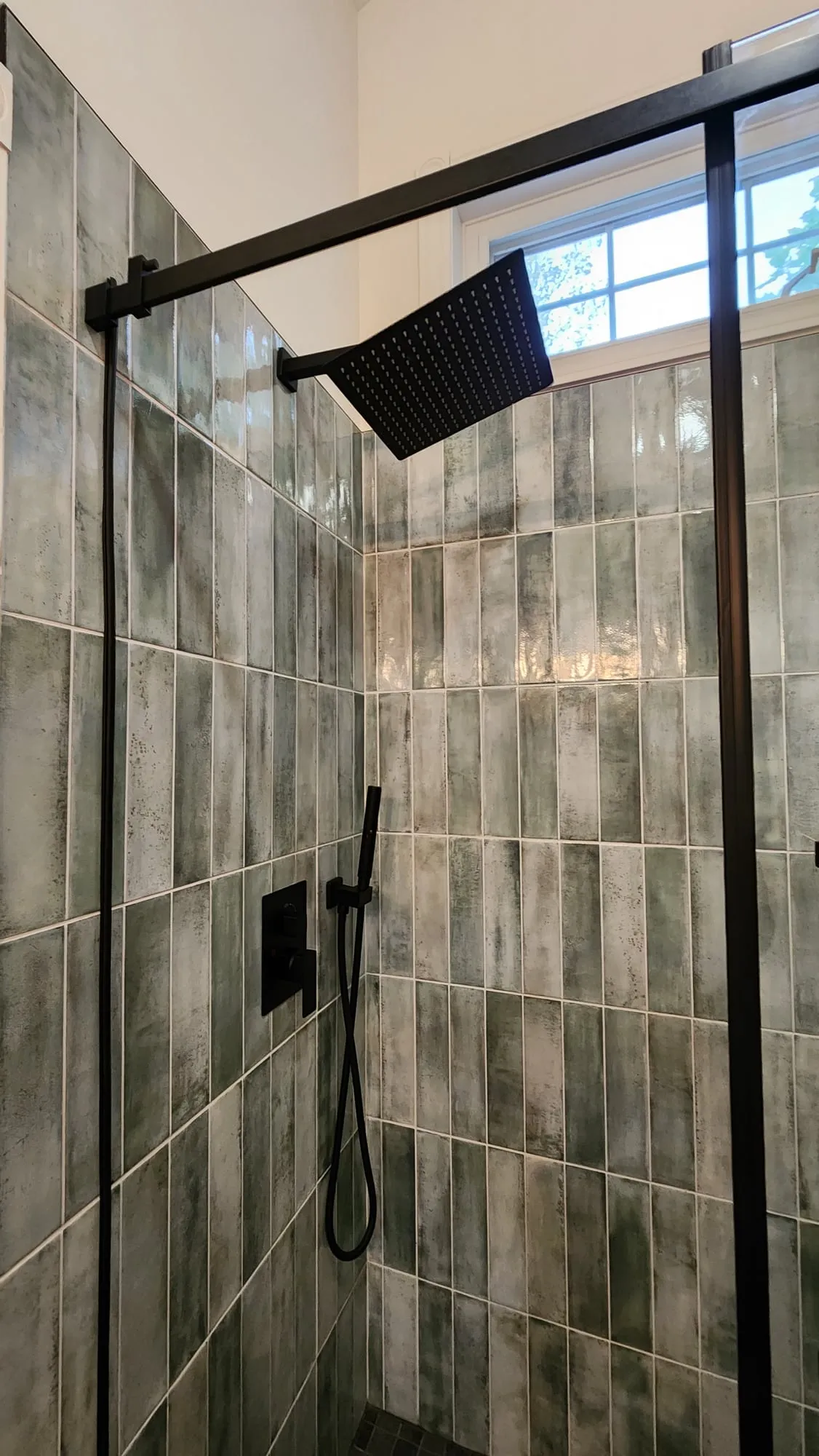 A close view of the shower with a black square rain head, handheld spray and vertical grey-green tile.