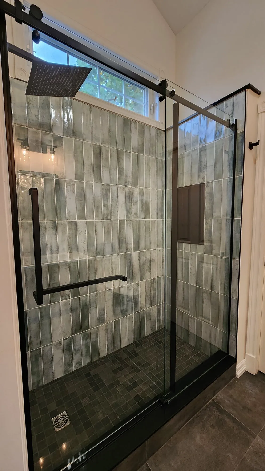 The finished shower behind a black-framed sliding glass door, with grey-green wall tile and a dark mosaic floor.