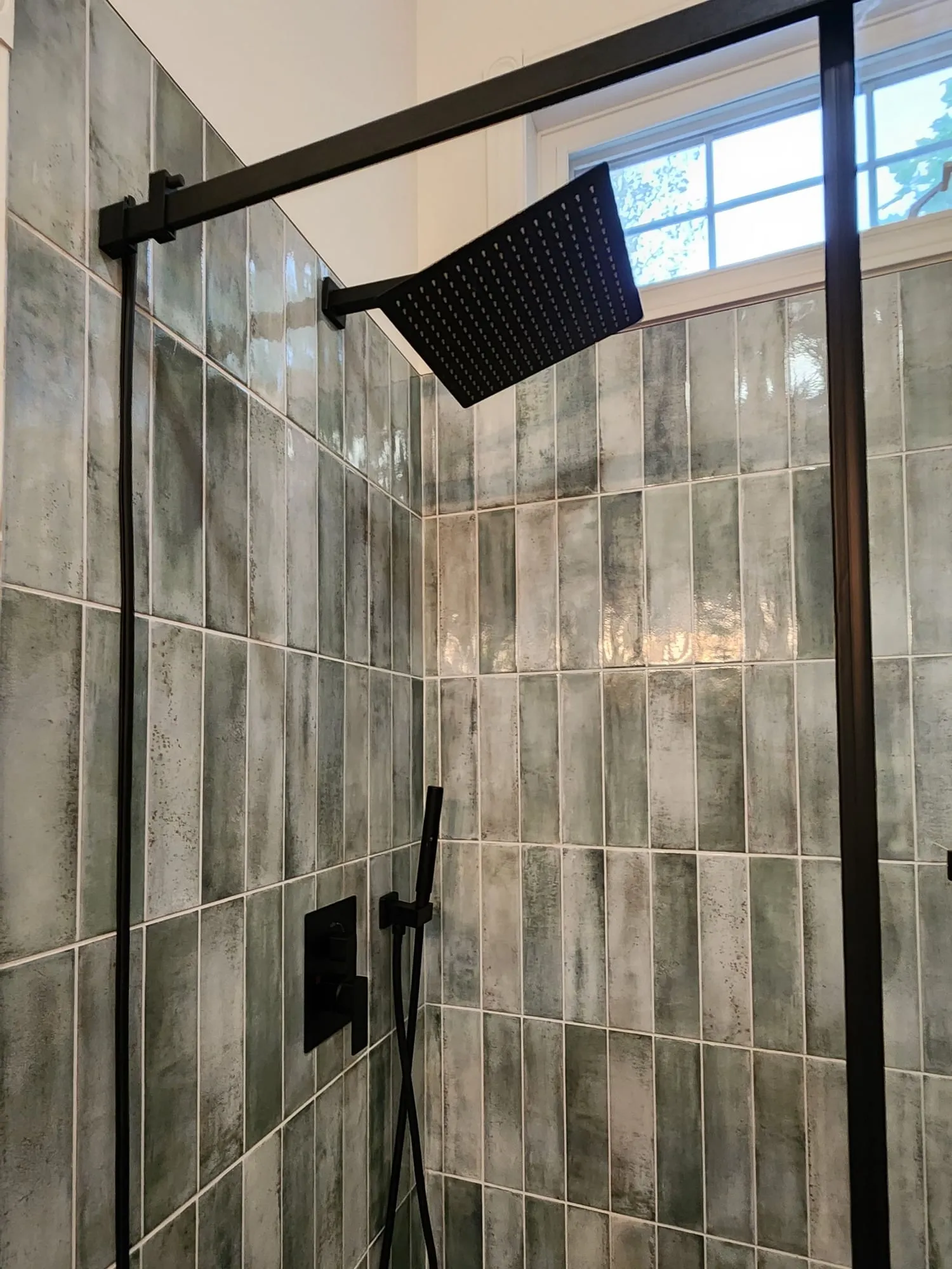A walk-in shower with green vertical tiles, a black rain shower head and a black-framed glass door