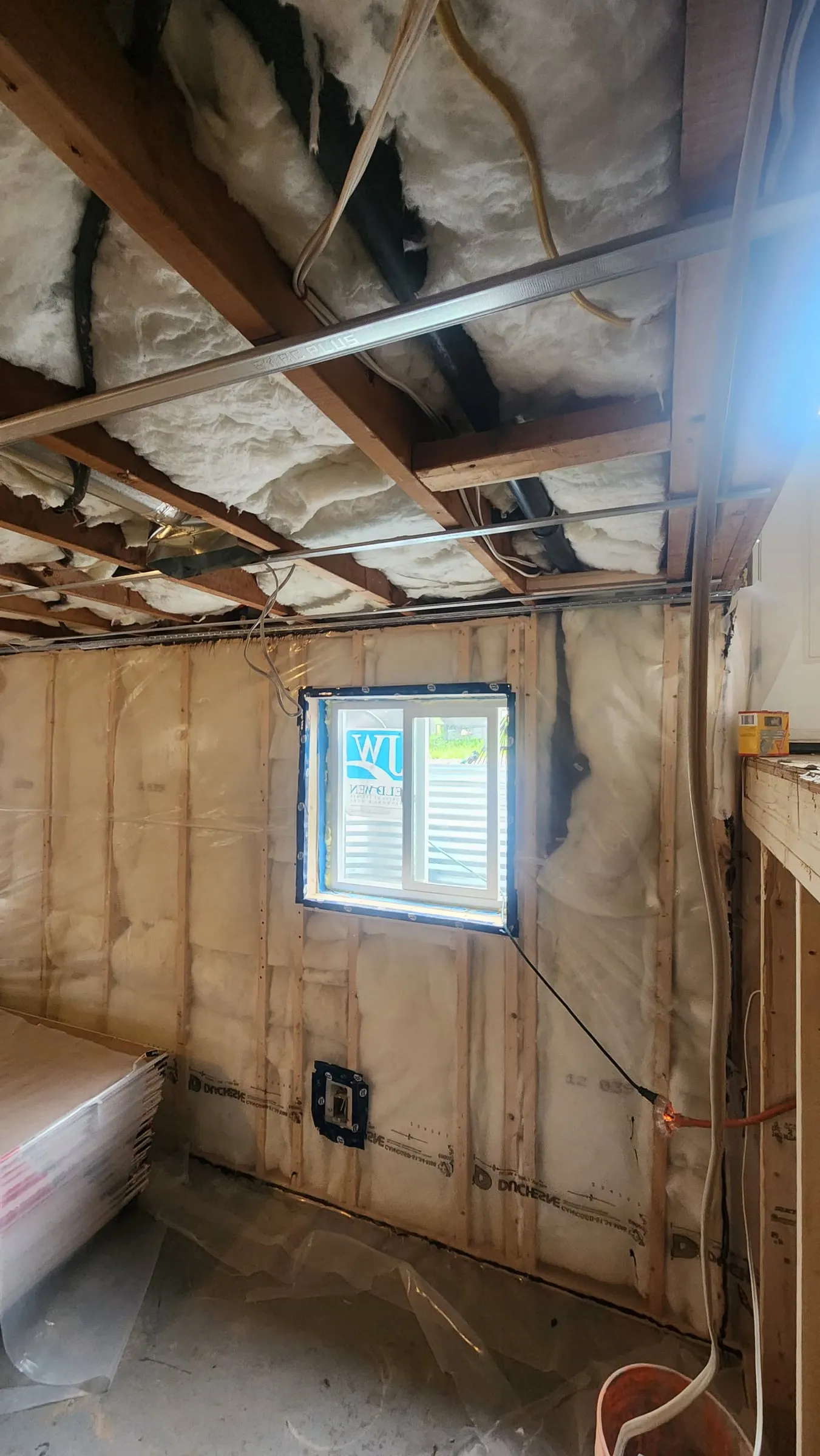 The bare basement: open ceiling joists with insulation, new walls framed and wrapped in plastic, and a small window.