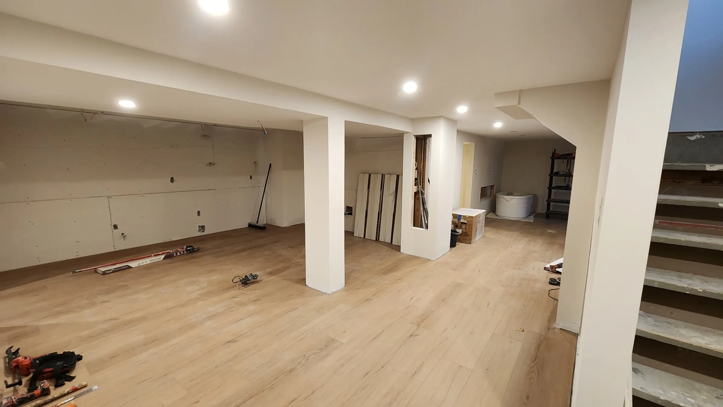 The open basement with new light wood-look flooring, white boxed-in posts, pot lights and the stairs on the right.
