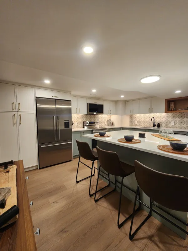 The basement kitchen: an island with three brown upholstered stools, sage-green lower cabinets, white uppers and a stainless fridge.