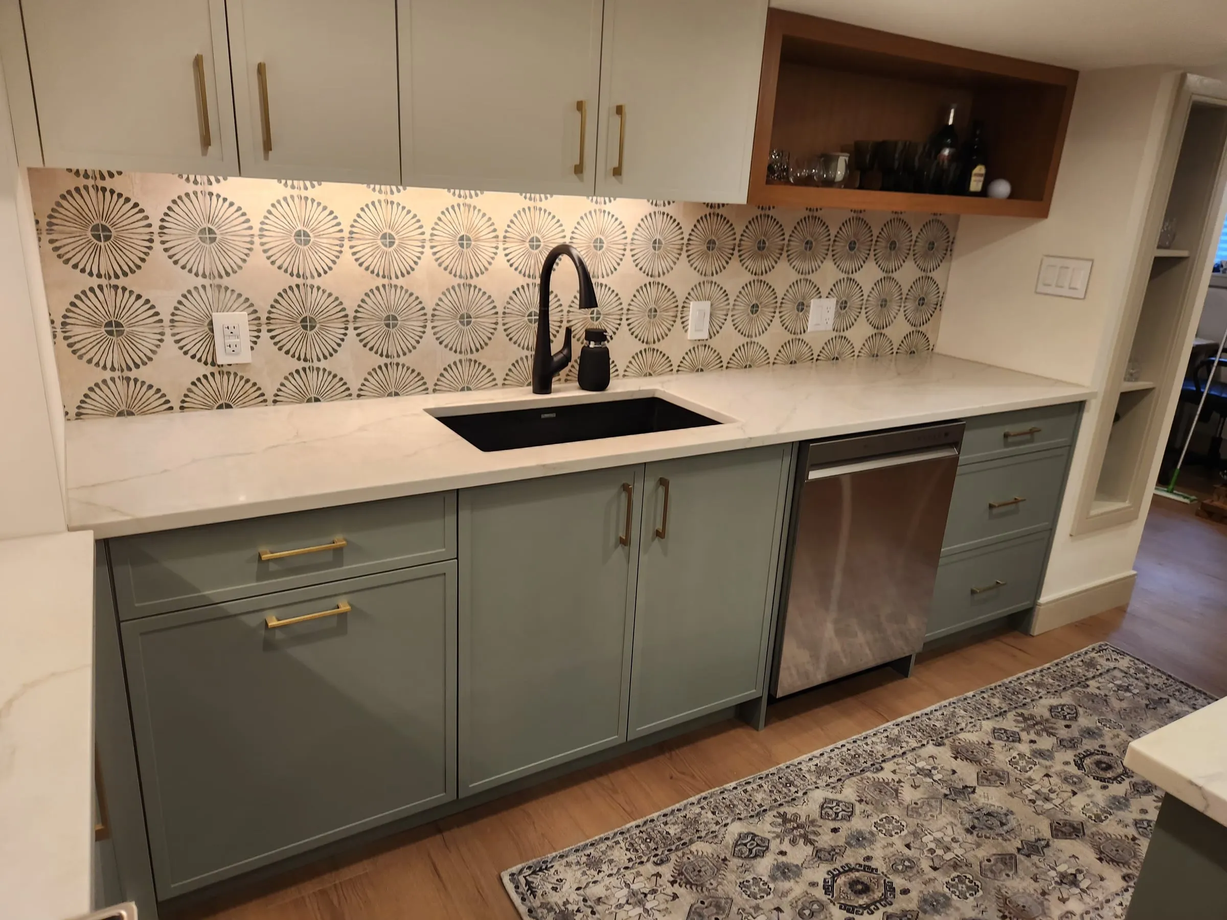 A black sink and faucet on a white counter, sage cabinets with brass pulls, a dishwasher and an open wood shelf above.