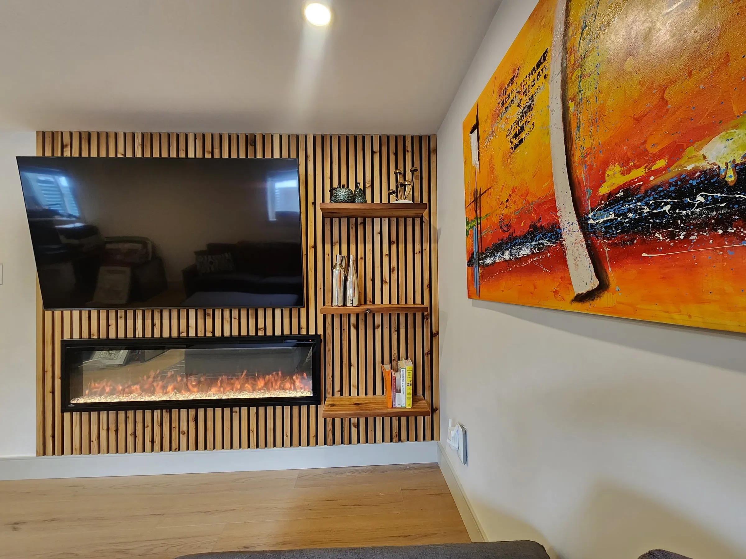 The slat wall up close, with floating wood shelves beside the TV and a bright orange painting on the side wall.