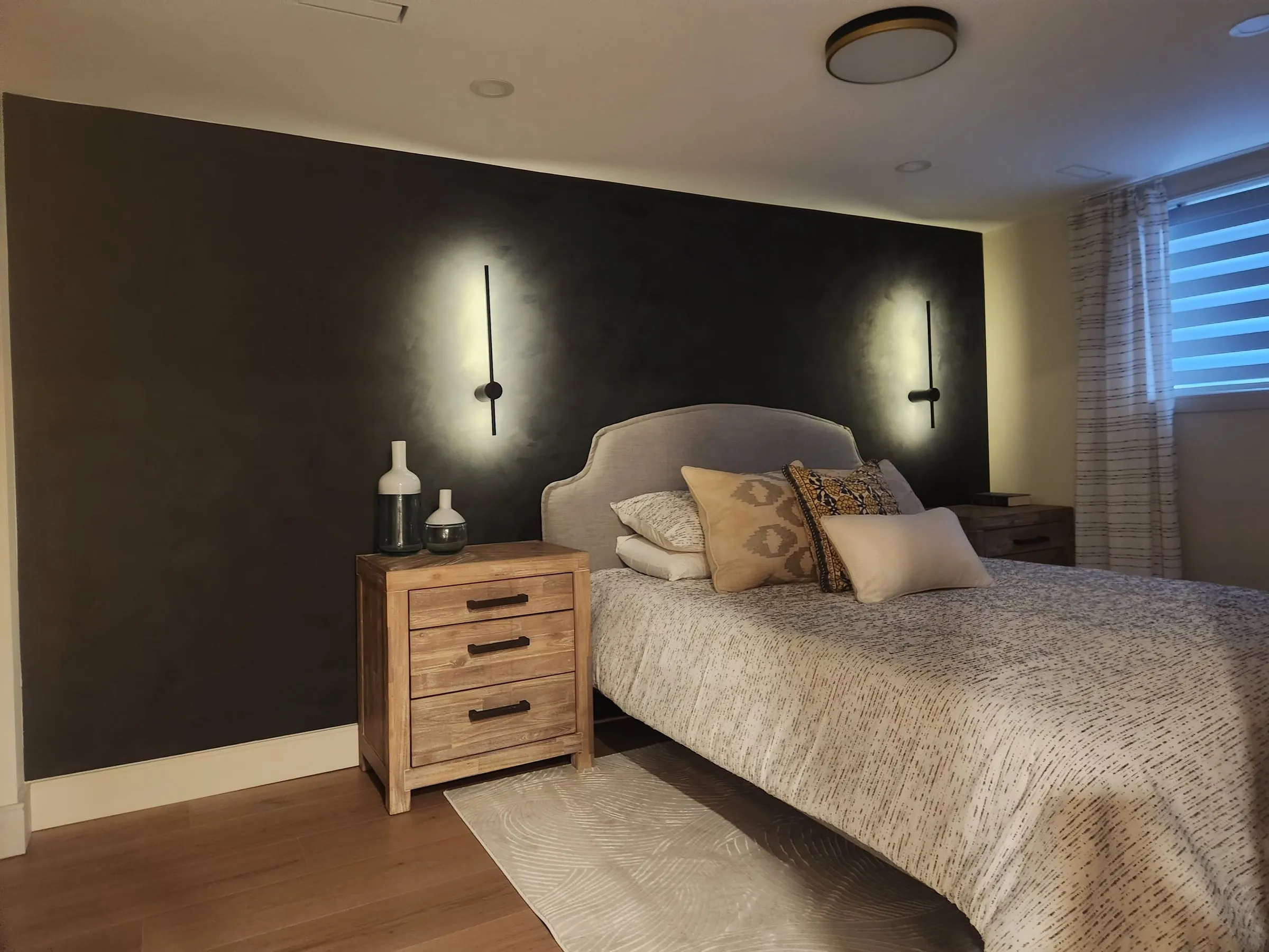 A bedroom with a dark accent wall, two slim black wall lights on either side of an upholstered bed, and wood nightstands.