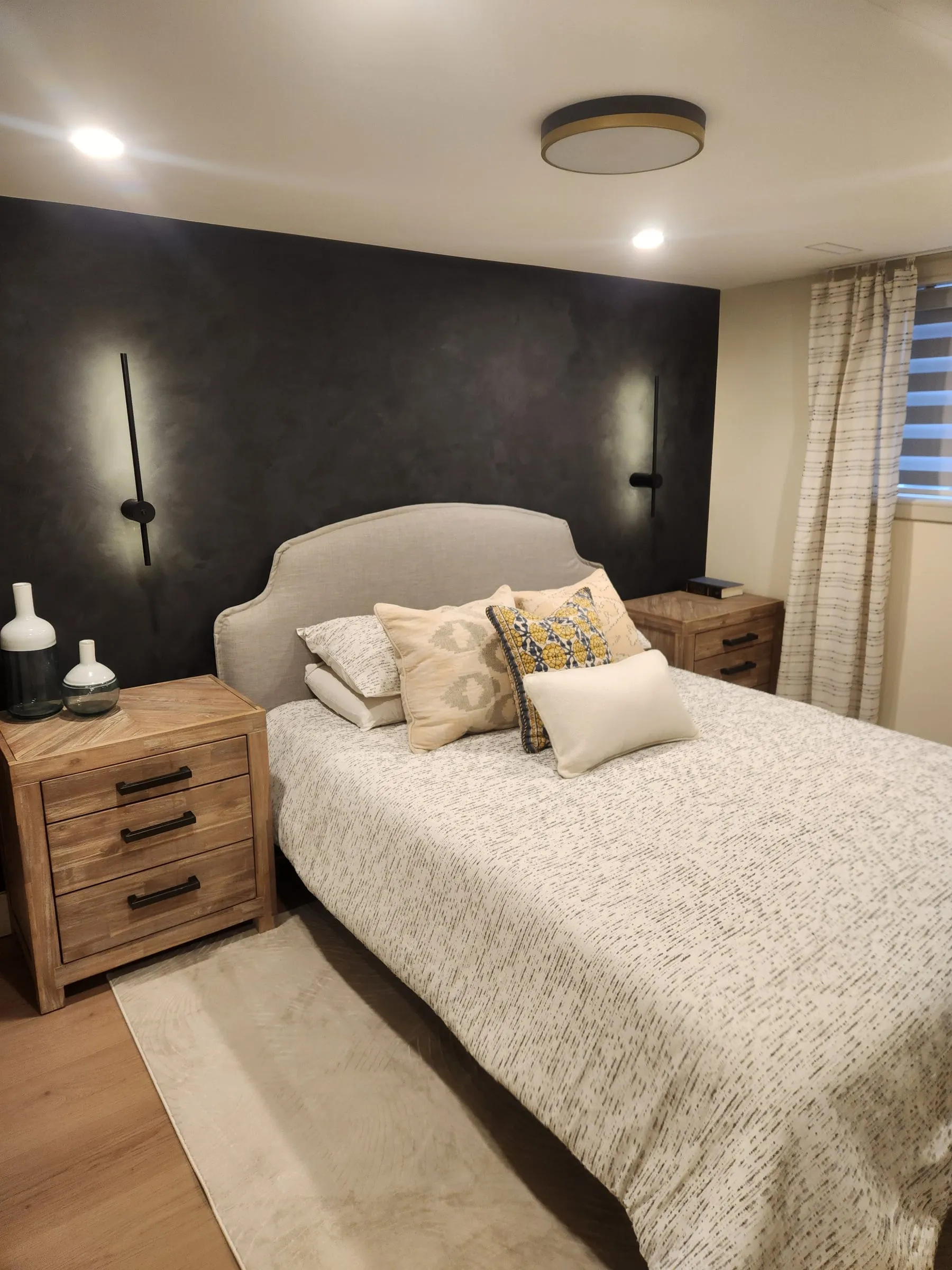 The bed against the dark accent wall, with a round ceiling light and a window with blinds.