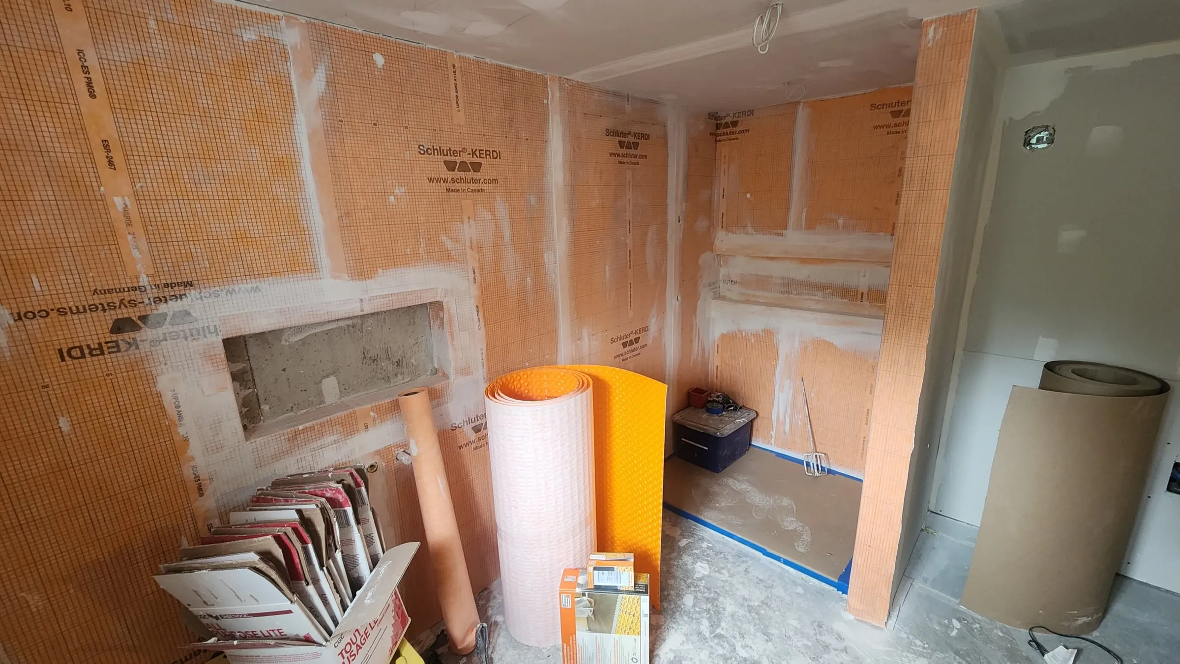 Orange waterproofing membrane on the bathroom walls, a recessed niche framed in and rolls of membrane on the floor.