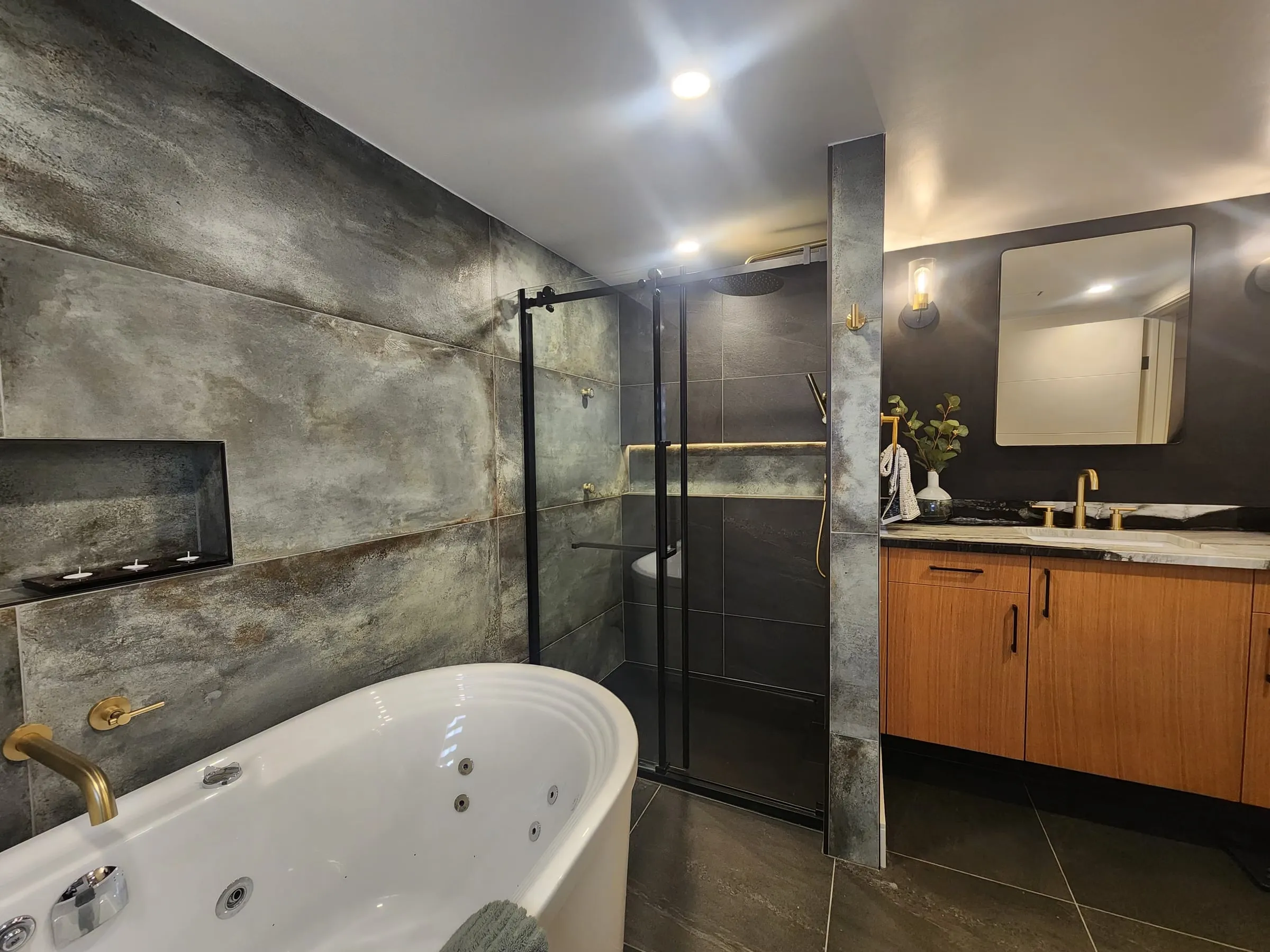 The finished bathroom: a white freestanding jetted tub, a glass shower with black frames, and a wood vanity under a mirror.