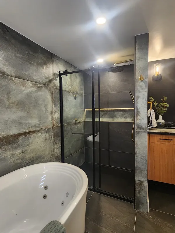 The finished bathroom: a white freestanding jetted tub, a glass shower with black frames, and a wood vanity under a mirror.