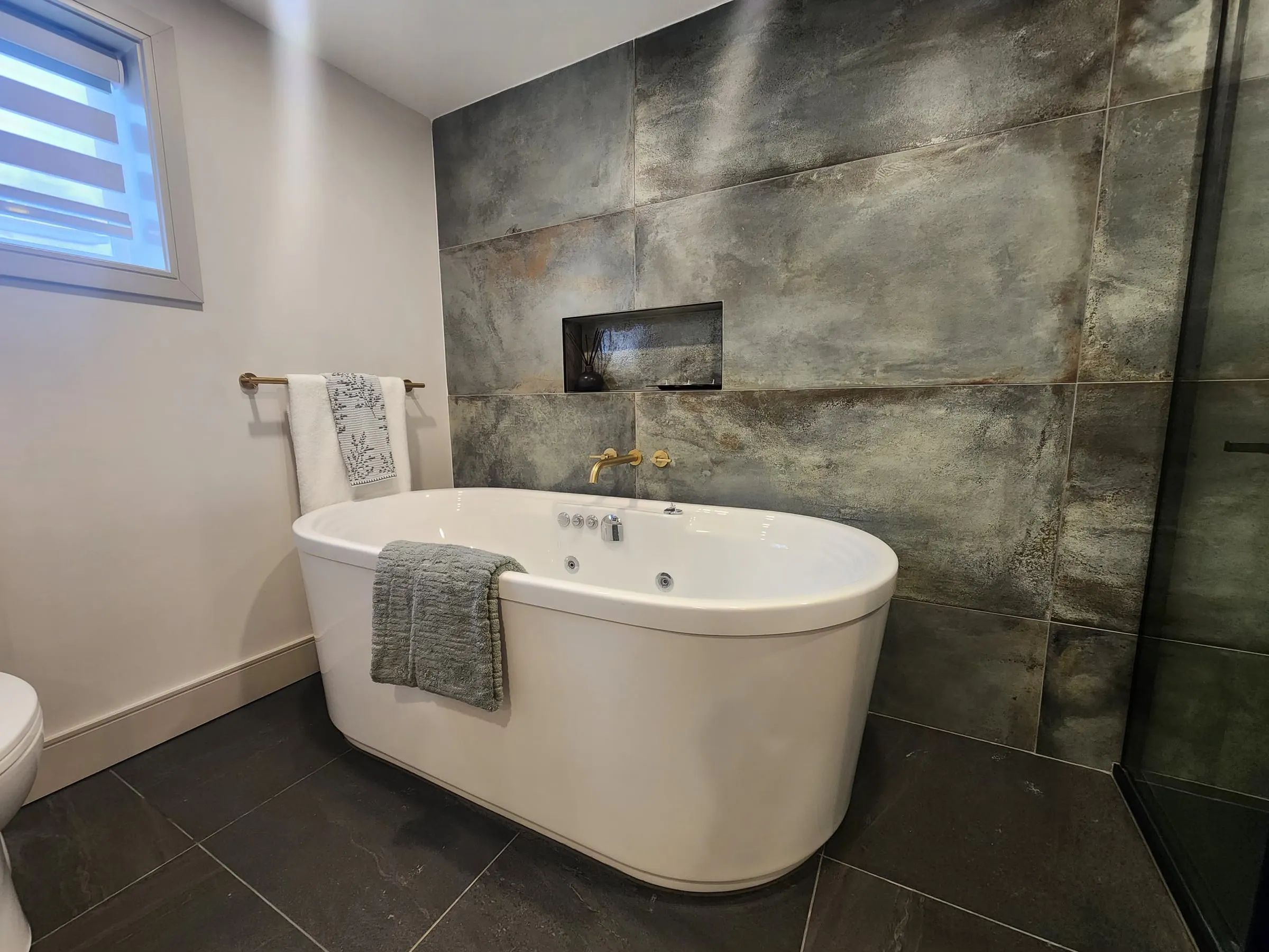 A white freestanding tub with jets against a wall of large grey-green tiles with a long niche.