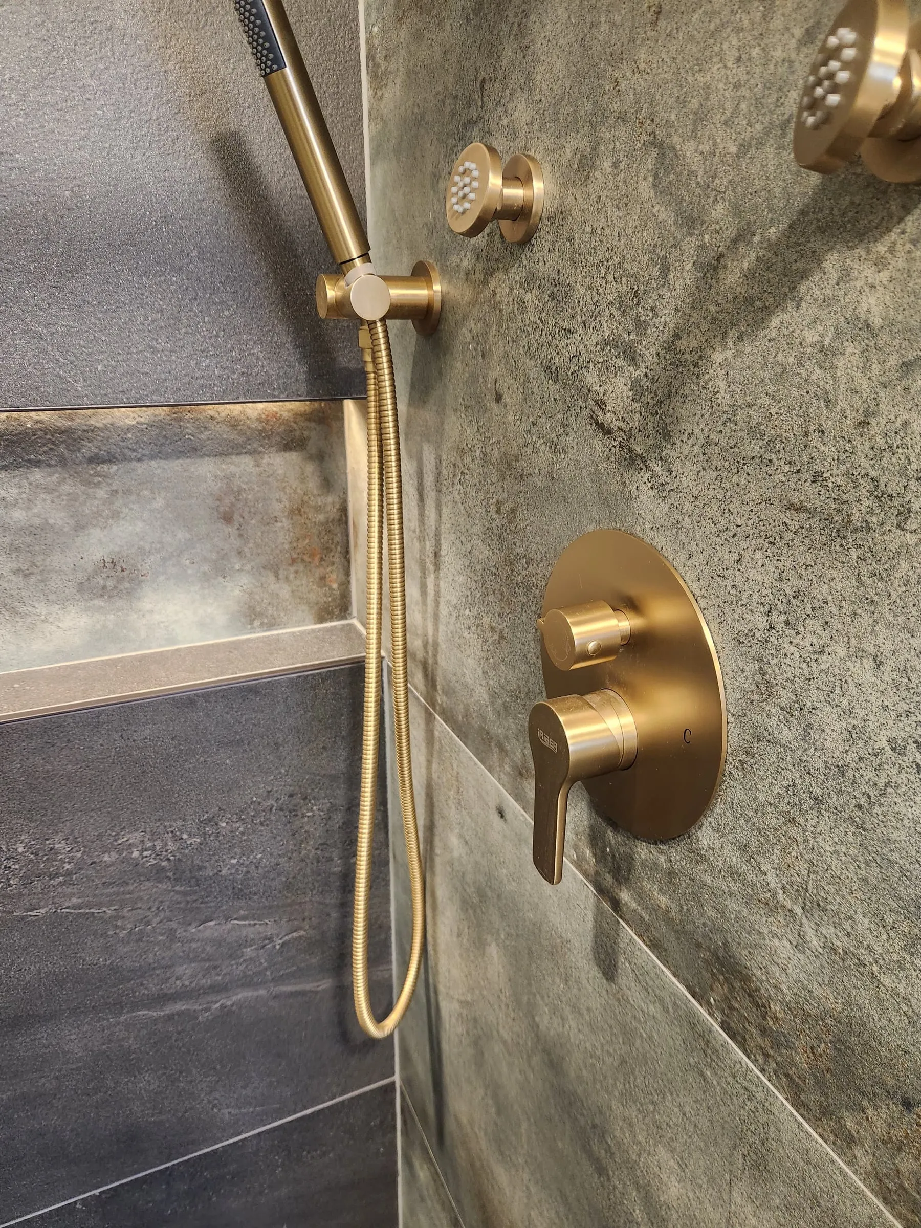 Brushed gold shower controls, a handheld spray on a hose and body jets on the tiled wall.