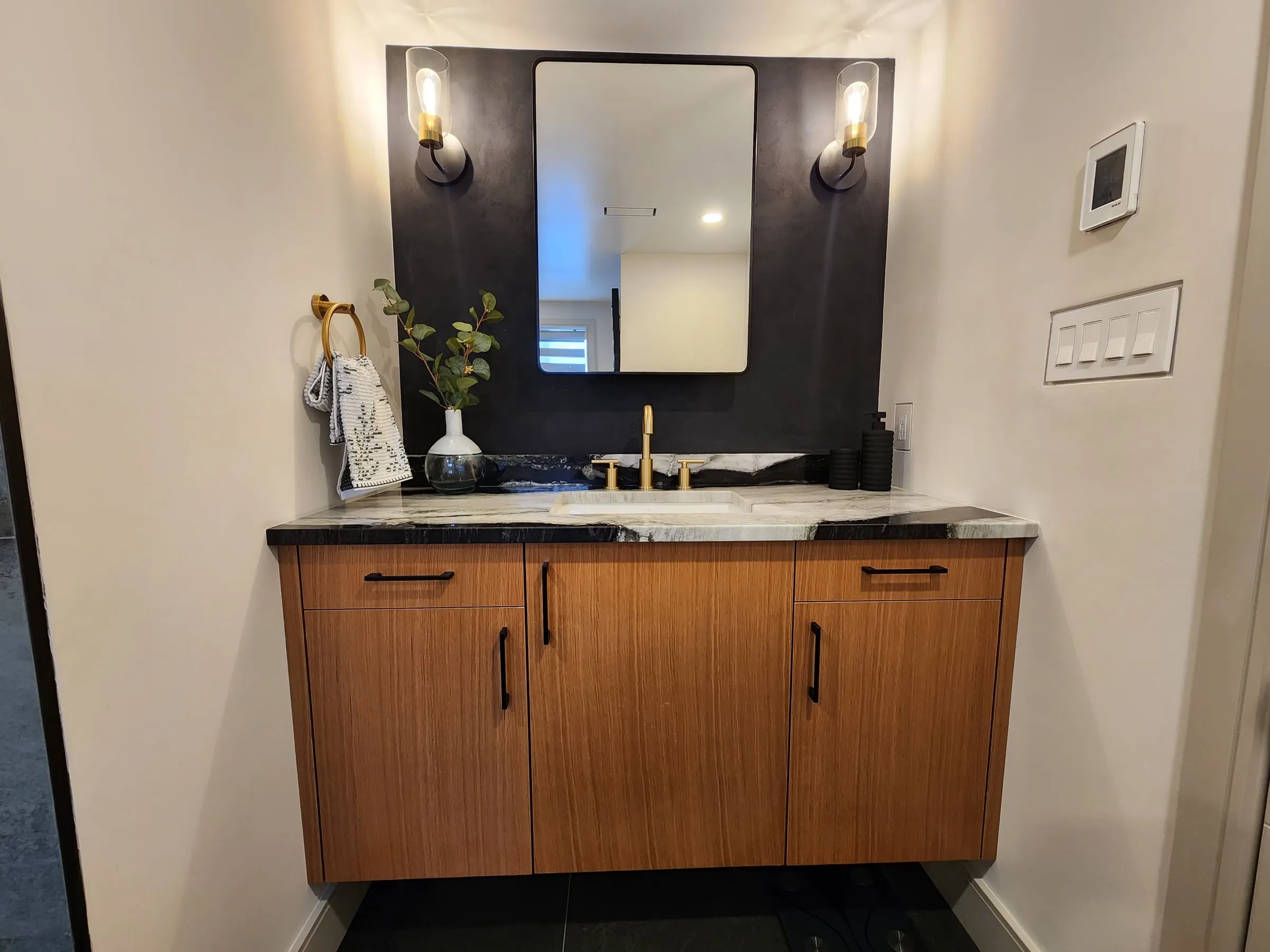 A floating wood vanity with a black and white veined top, against a black wall with a mirror and two wall lights.