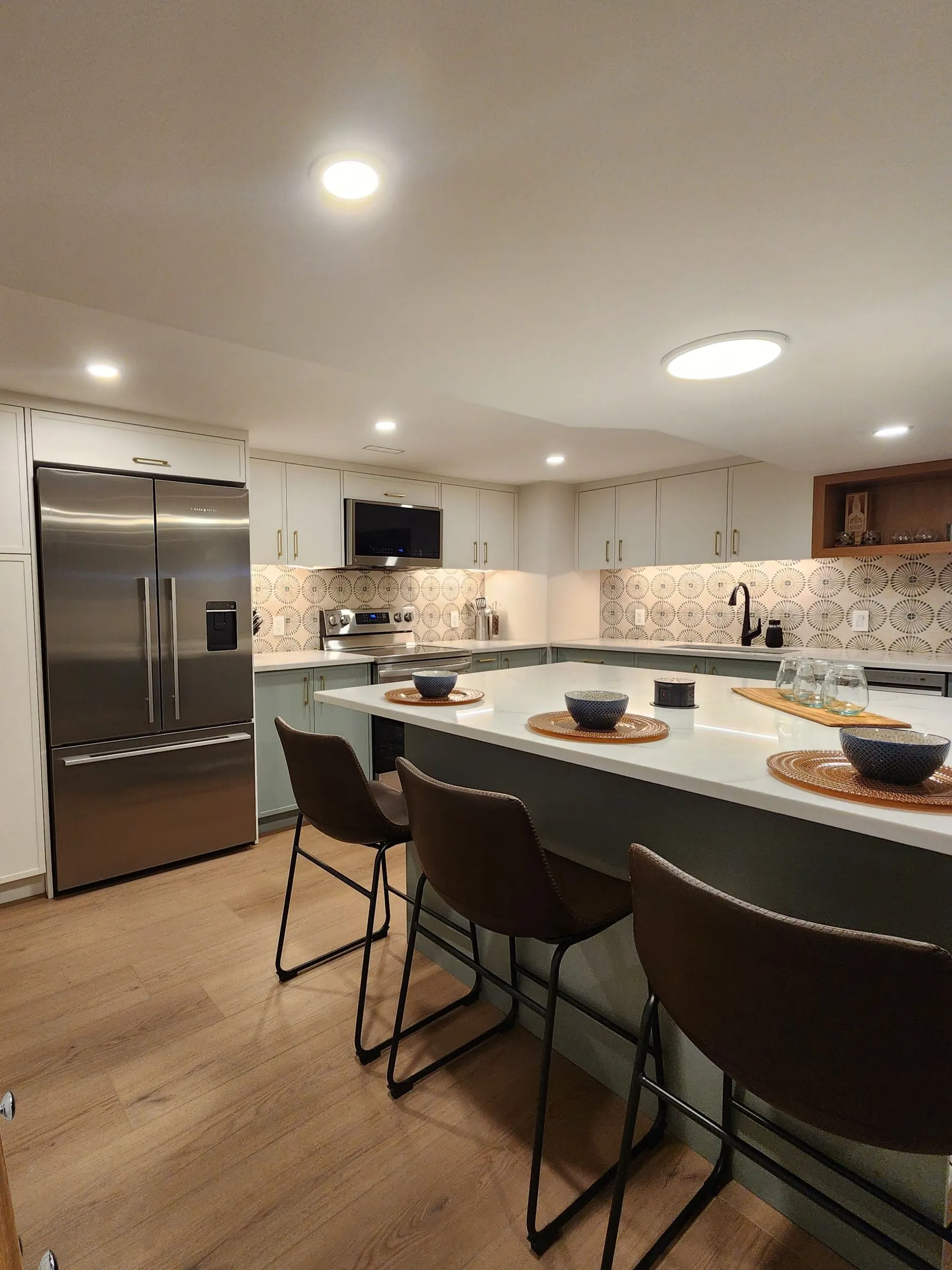 A basement kitchen with sage lower cabinets, white uppers, an island with three brown stools and a stainless fridge