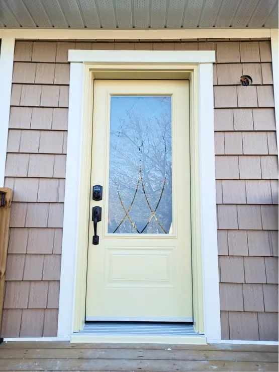 A new pale yellow front door with a decorative glass panel, black hardware and tan shingle siding.