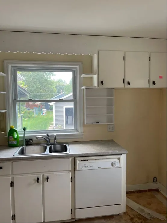 The old kitchen, with plain white cabinets, a grey countertop, a double sink and a white dishwasher.