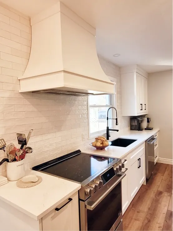 The finished kitchen: a white range hood over the stove, white tile backsplash, black sink and white cabinets.