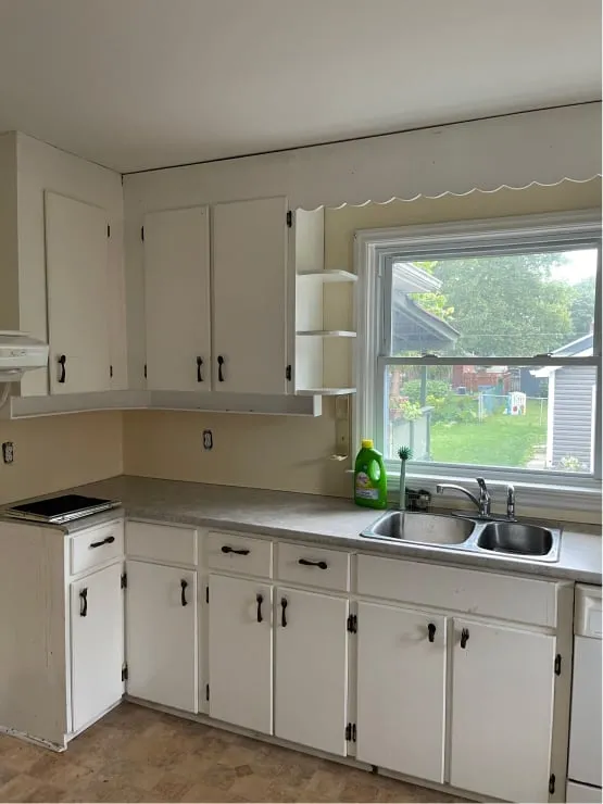 The old kitchen before, with white cabinets, a scalloped valance over the window and a patterned floor.