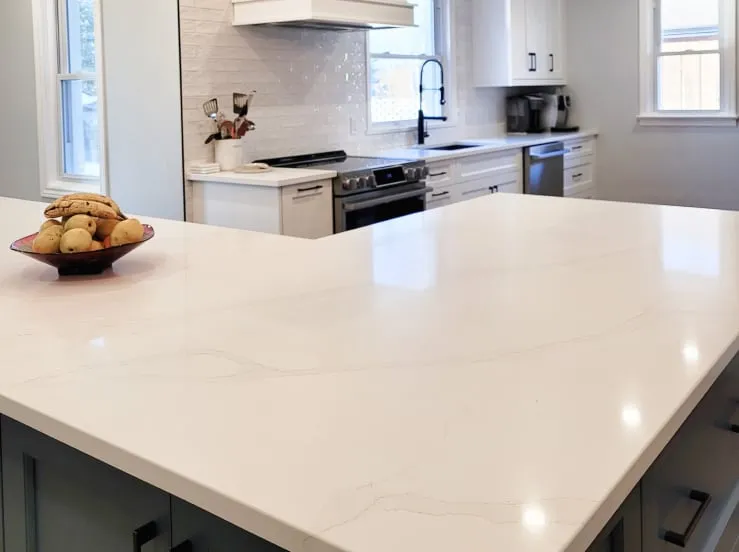 A wide white island countertop with soft grey veins, looking toward the finished stove wall and windows.