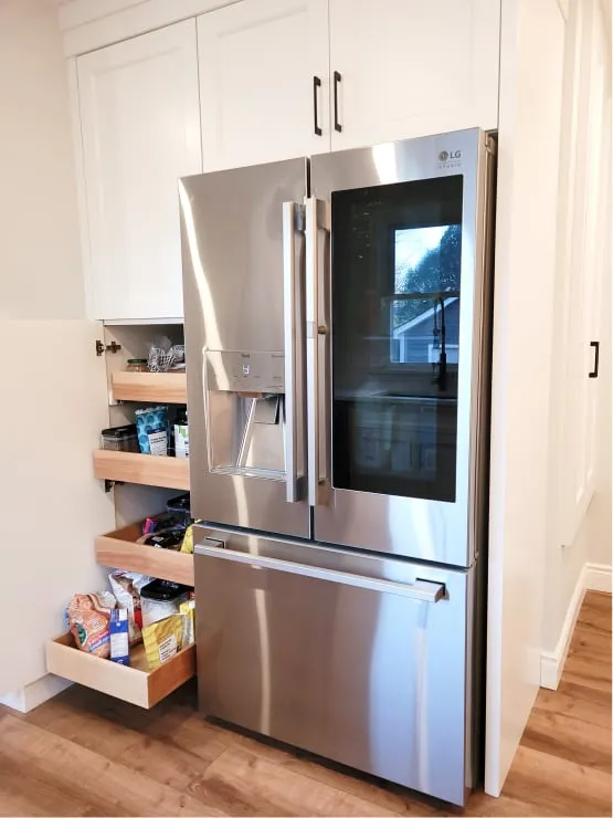 Pull-out wood shelves stocked with food inside a tall white cabinet beside the silver fridge.