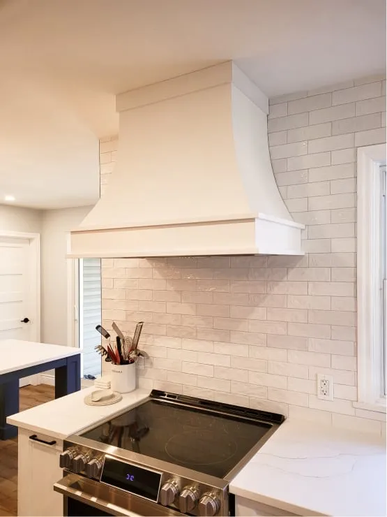 A white range hood over a black smooth-top stove and glossy white wall tile, with the blue island at left.