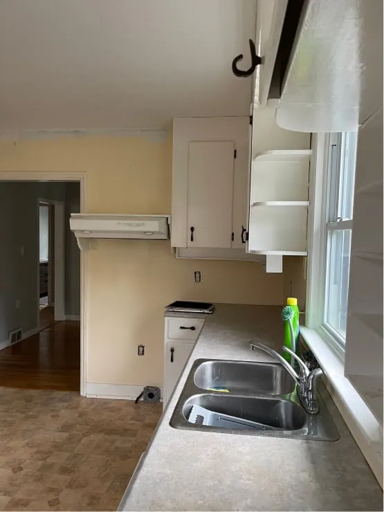 The old sink corner before, with a double basin, grey countertop, open white shelves and a patterned floor.