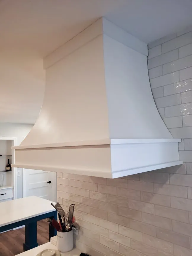 The curved white range hood up close, against glossy white wall tiles with a utensil holder below.