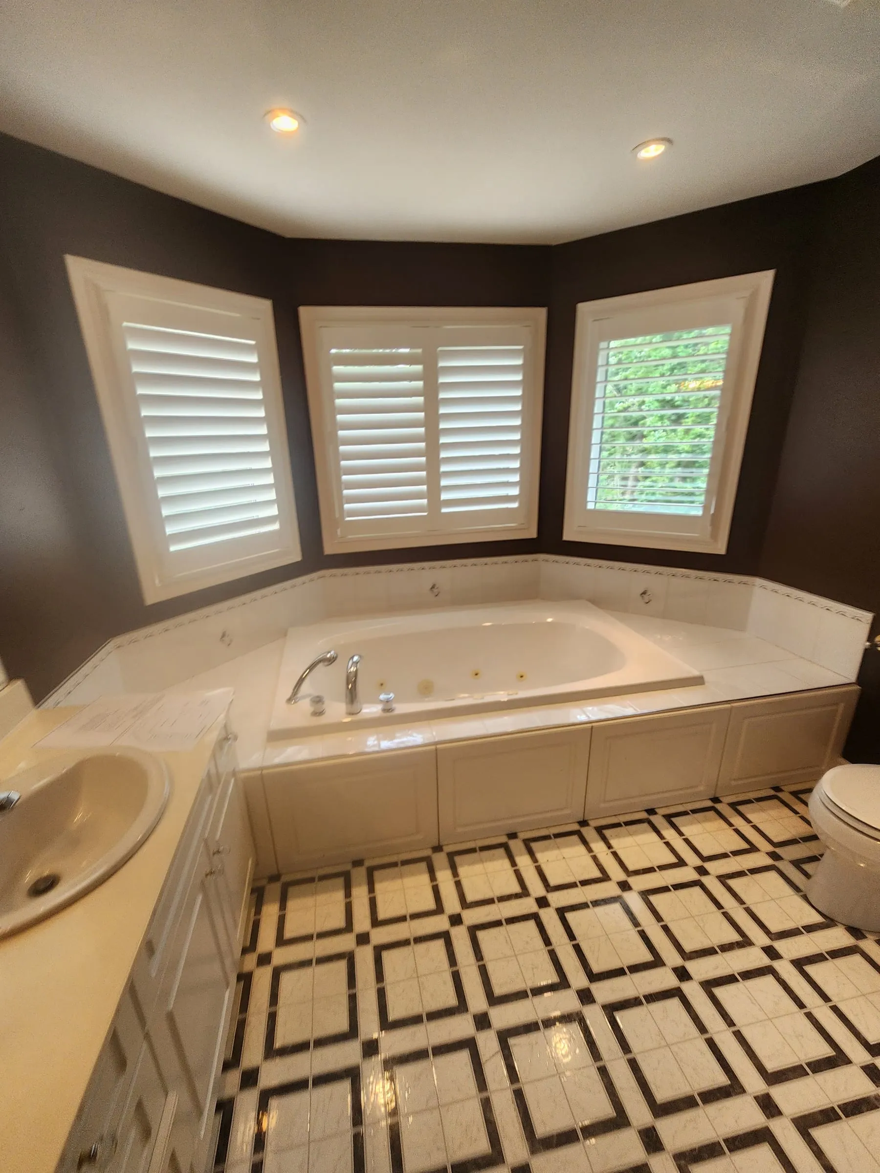 The old bathroom: a jetted tub set in a tiled deck under three shuttered windows, dark brown walls and a black and white patterned tile floor.