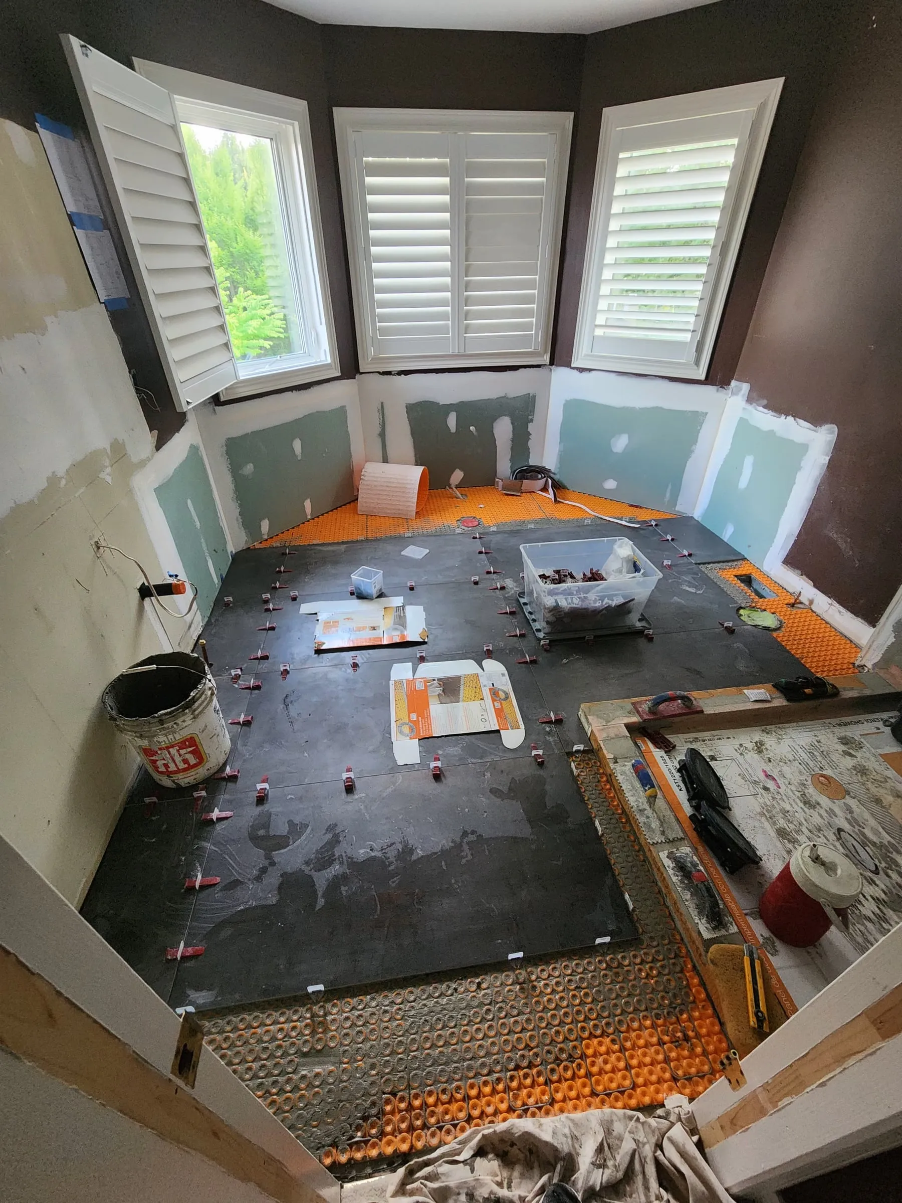 Large dark floor tiles being laid with red levelling clips across the room, under the bay of shuttered windows.
