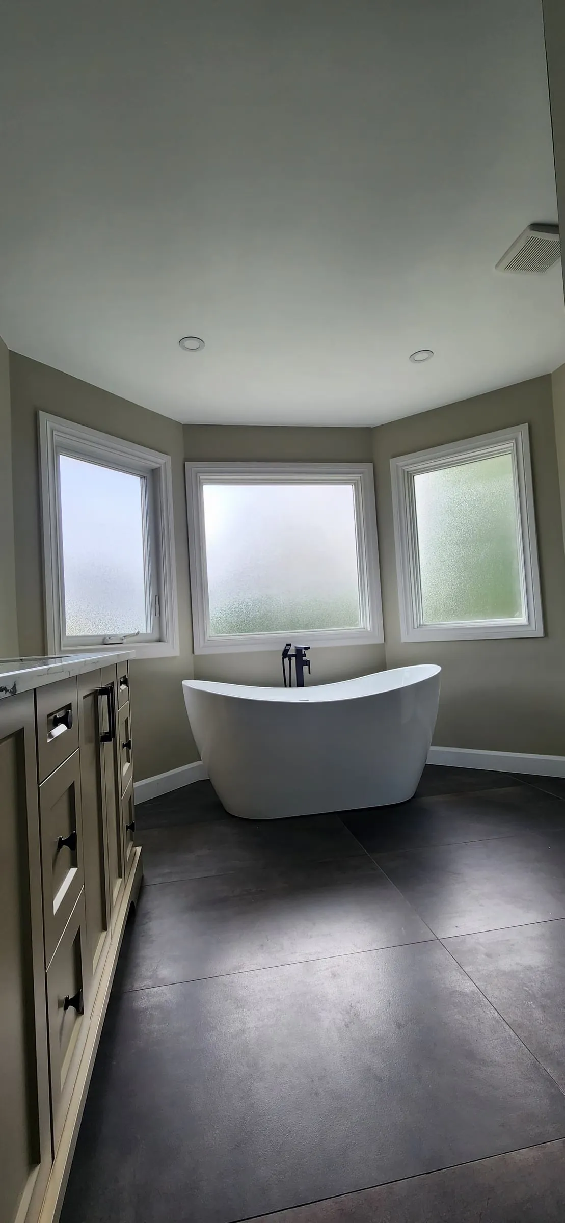 The finished bathroom: a white freestanding tub in the bay of three frosted windows, a dark tile floor and the vanity on the left.