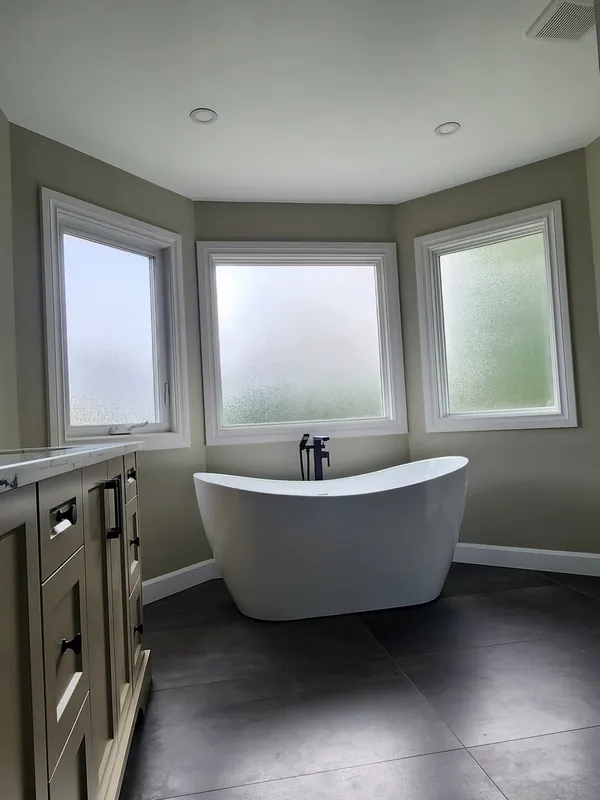 The finished bathroom: a white freestanding tub in the bay of three frosted windows, a dark tile floor and the vanity on the left.
