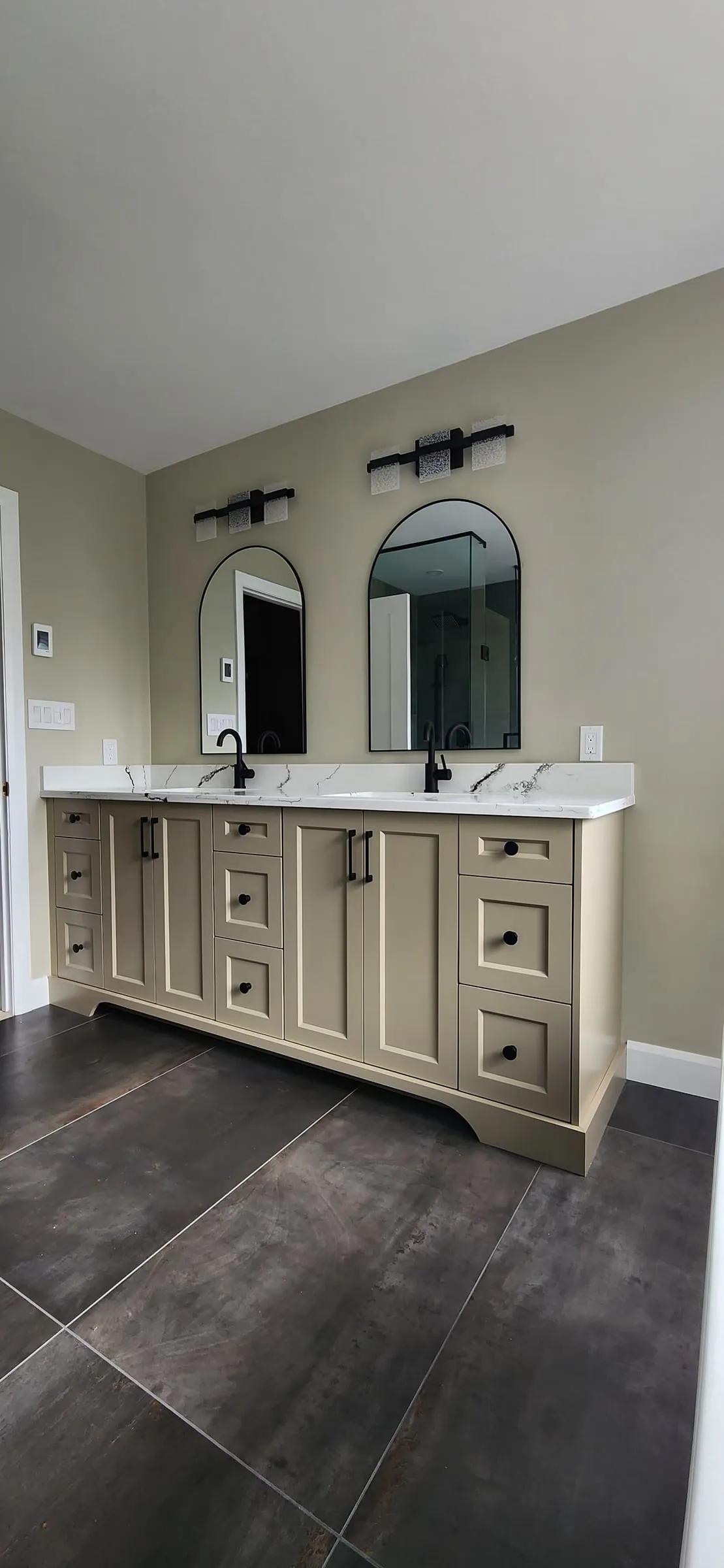 A double vanity with beige shaker cabinets, a white top, two black faucets and two arched black mirrors.