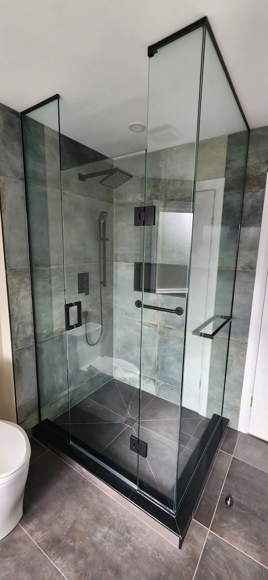 A glass shower with black hardware, grey-green tile walls, a rain head and a dark shower base.