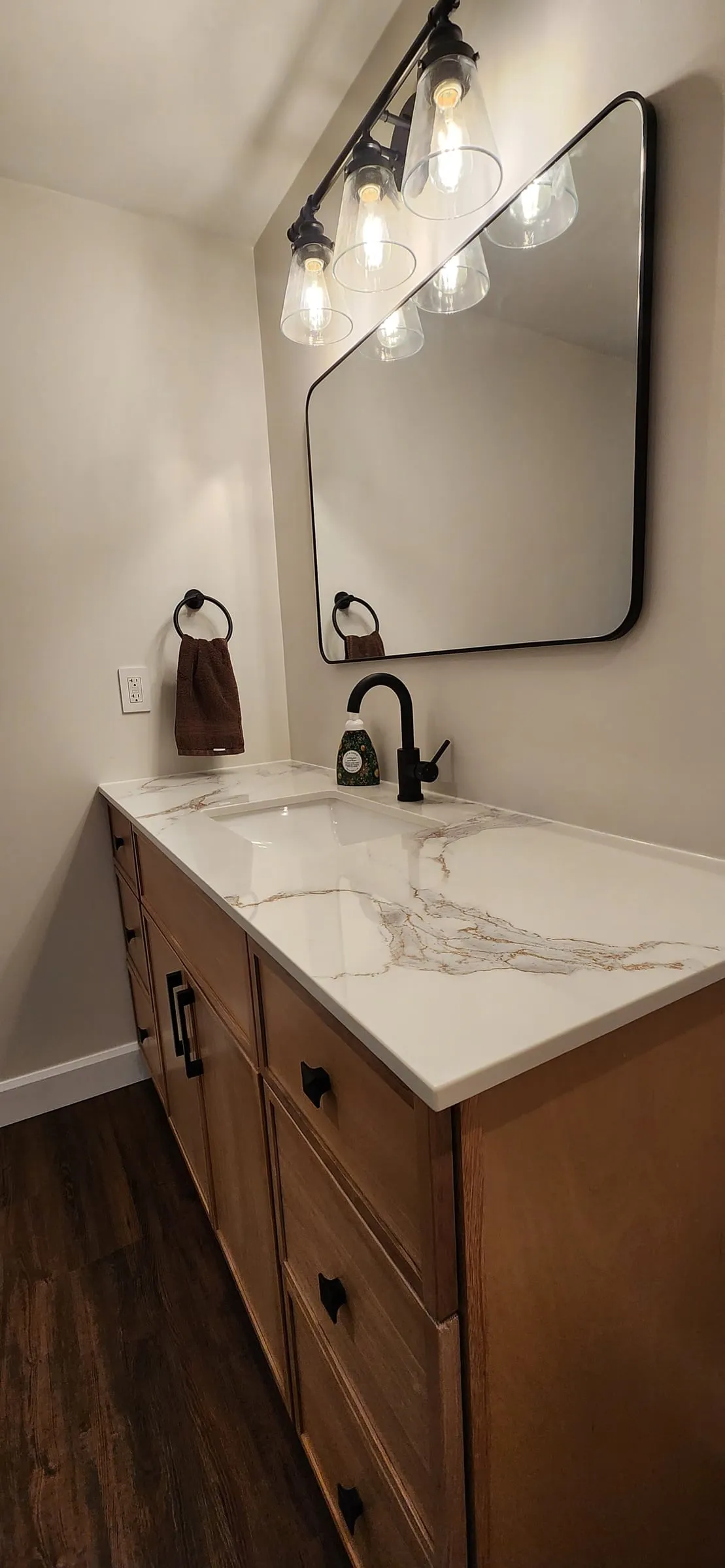 A wood vanity with a veined white top, a black faucet, a black-framed mirror and a light with clear glass shades.