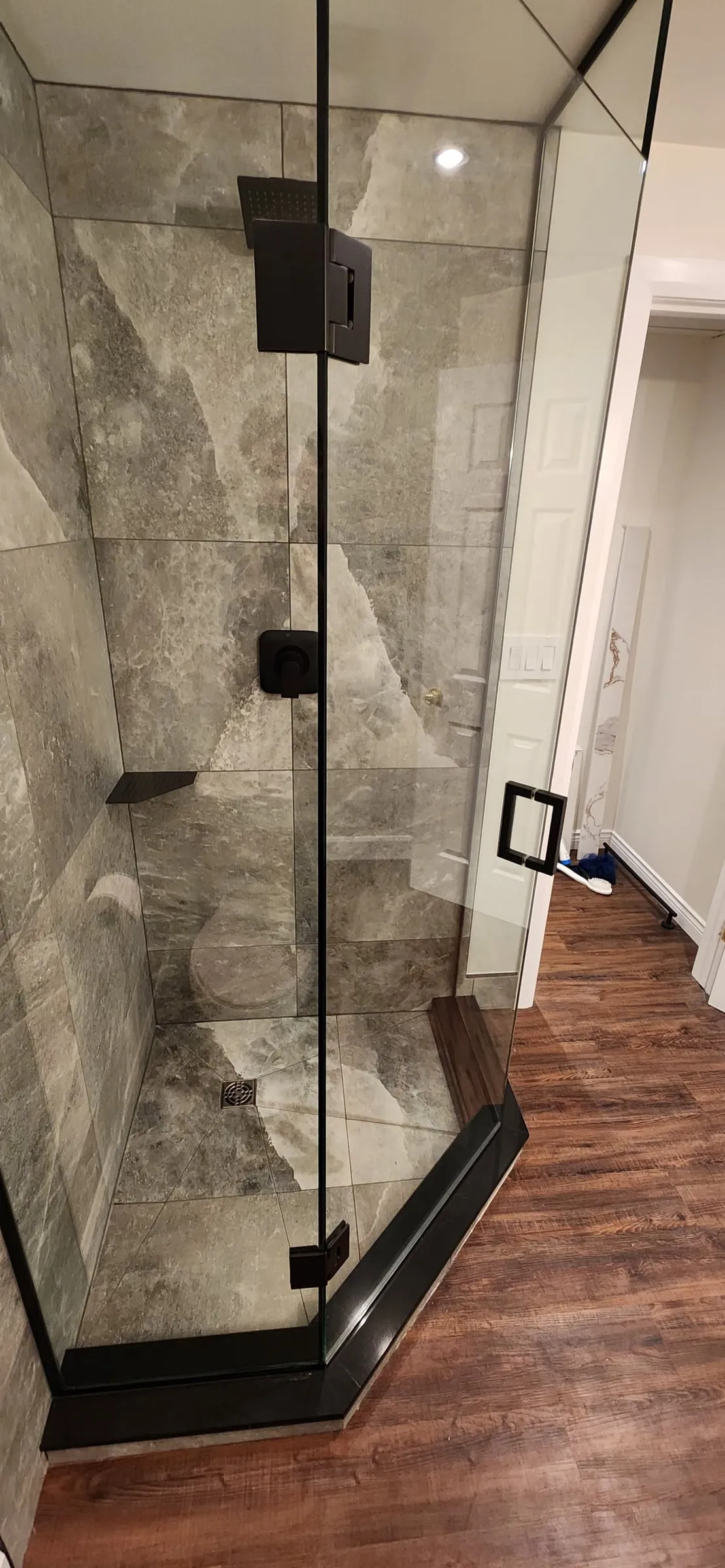 A corner glass shower with black hardware and grey stone-look tile, beside a wood-look floor.