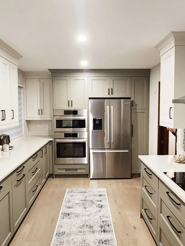 A long kitchen with sage lower cabinets, white and sage upper cabinets, stacked wall ovens and a stainless fridge