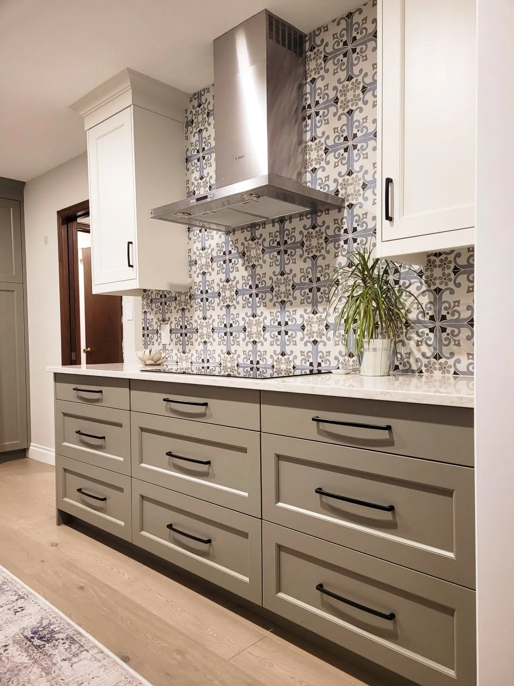Sage green kitchen drawers under a white veined countertop, a patterned blue tile backsplash and a stainless range hood
