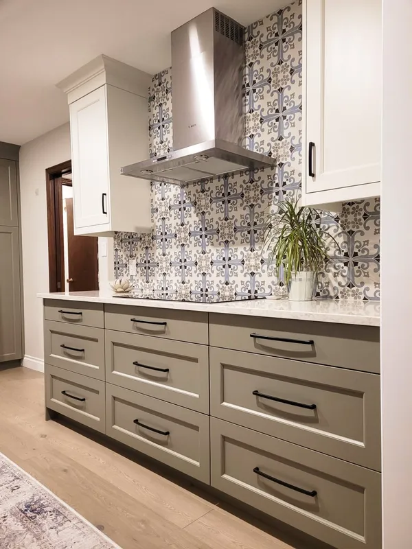 The finished kitchen with sage green drawers, white upper cabinets, a patterned blue-grey backsplash and metal range hood.
