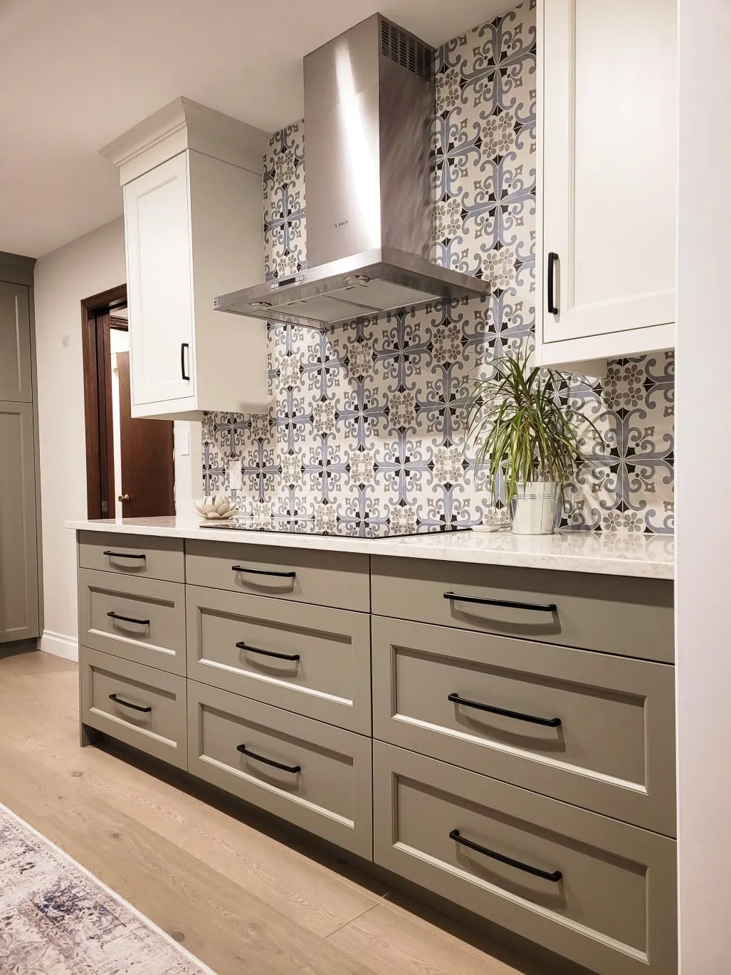 The finished kitchen with sage green drawers, white upper cabinets, a patterned blue-grey backsplash and metal range hood.