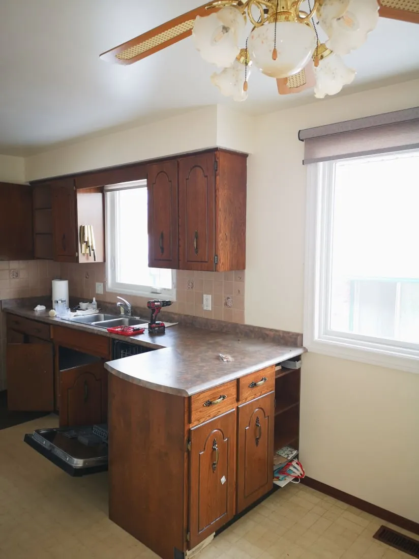 The old kitchen before the work, with an L-shaped speckled brown counter, wood cabinets, a double sink and ceiling fan.
