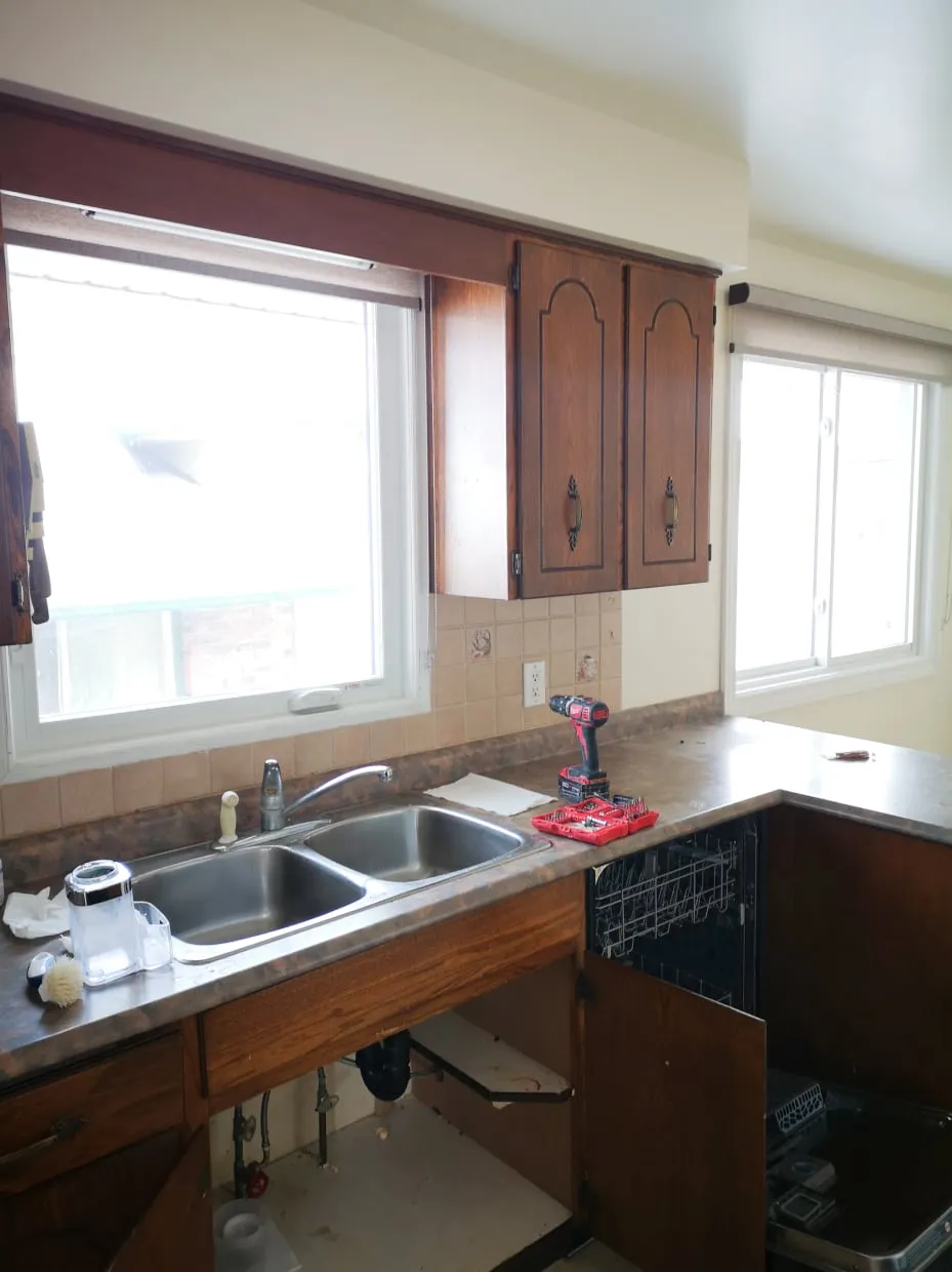 The old sink wall before the work, with a double sink, open cabinets, exposed pipes and two windows.