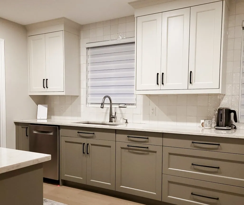 The finished sink wall with sage green lower cabinets, black pulls, white wall tile and a window shade.