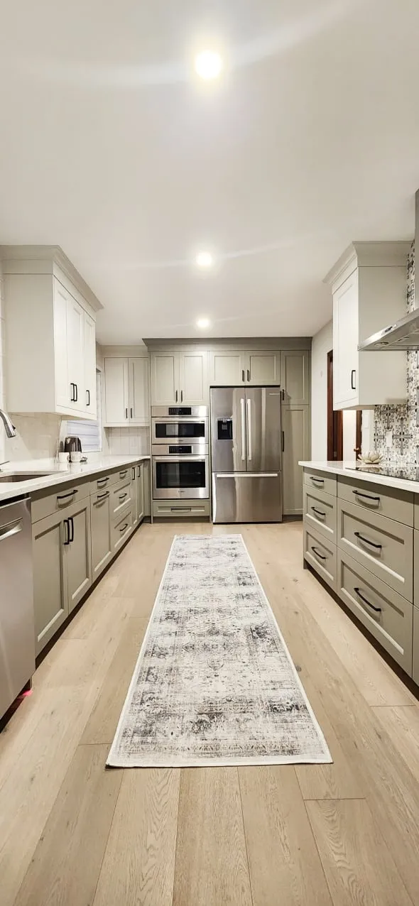 The finished kitchen seen down its centre, with sage green cabinets, stacked wall ovens, a fridge and rug.