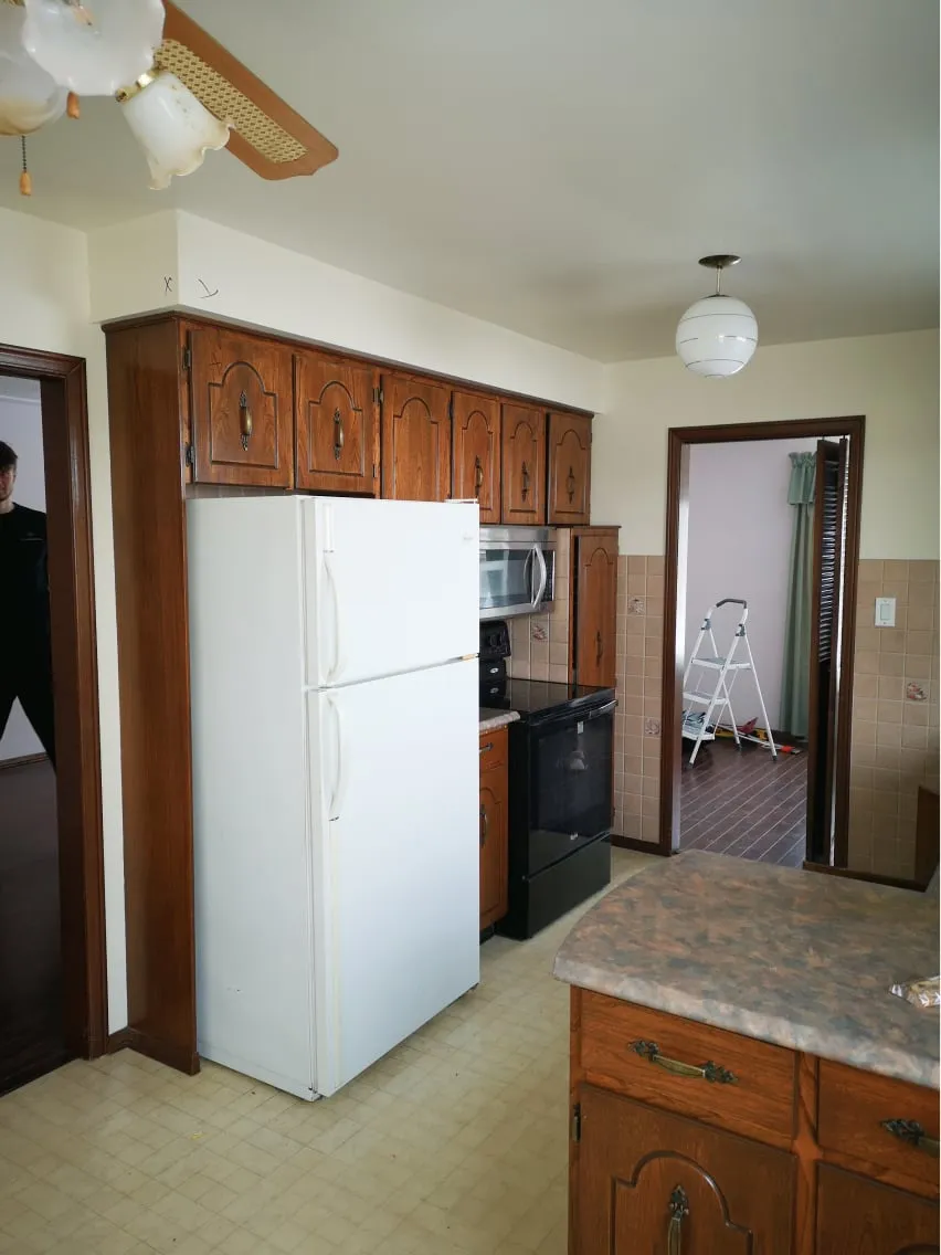 The old kitchen before the work, with a white fridge, wood cabinets, a black stove and narrow doorway.