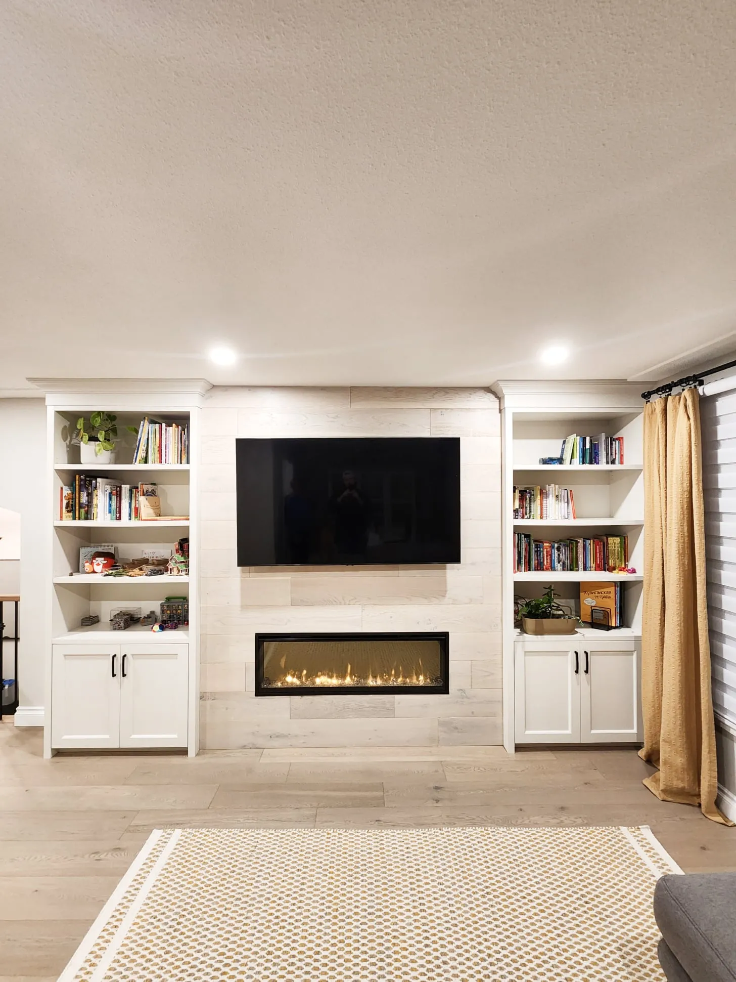 The finished living room wall with a fireplace and TV on pale plank panelling between white built-in bookshelves.