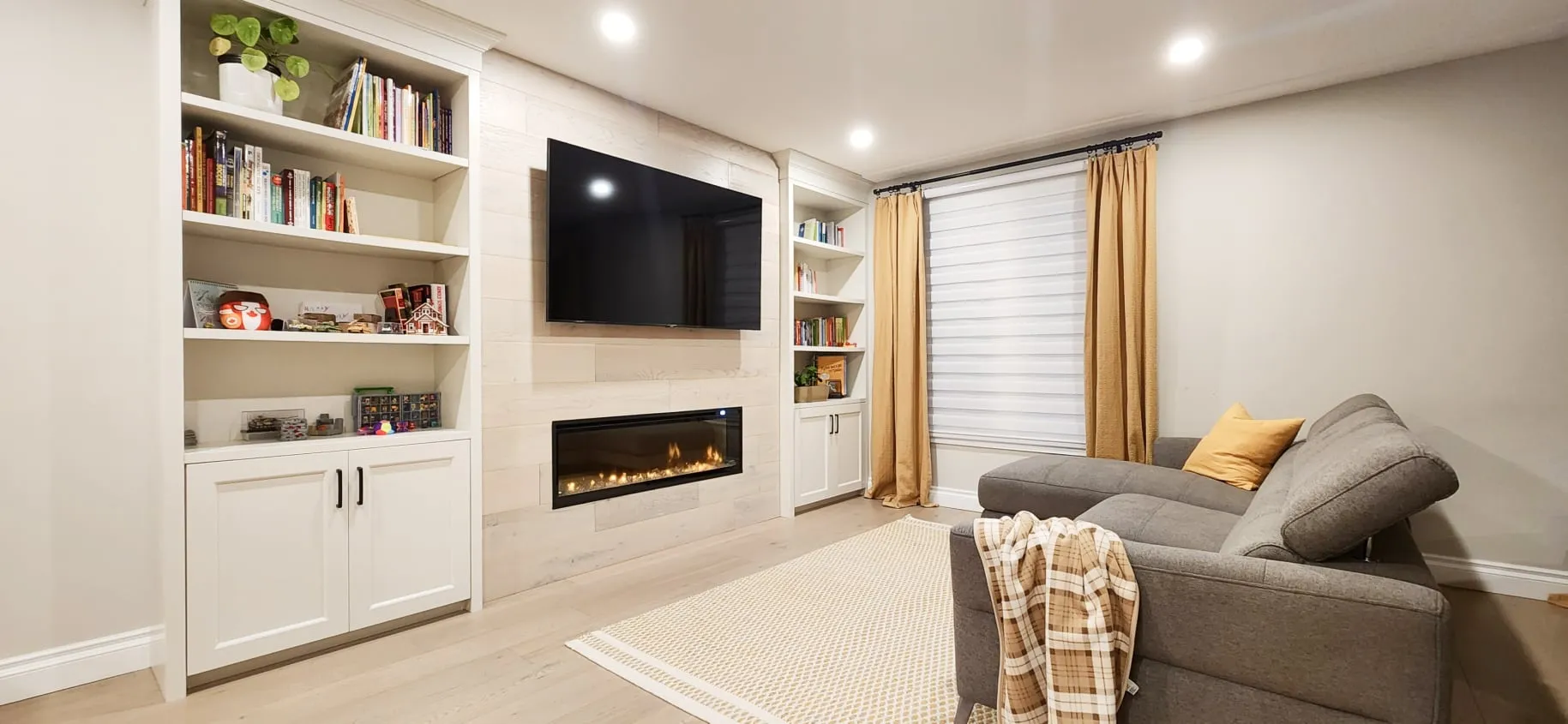 The finished living room with a grey sectional sofa facing the TV, fireplace and white built-in shelves.