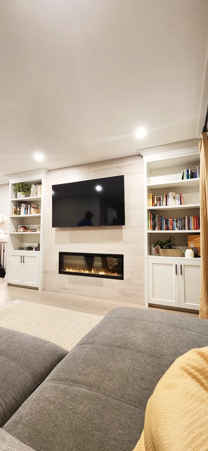 The finished living room seen from the grey sofa, facing the lit fireplace, TV and white built-in shelves.