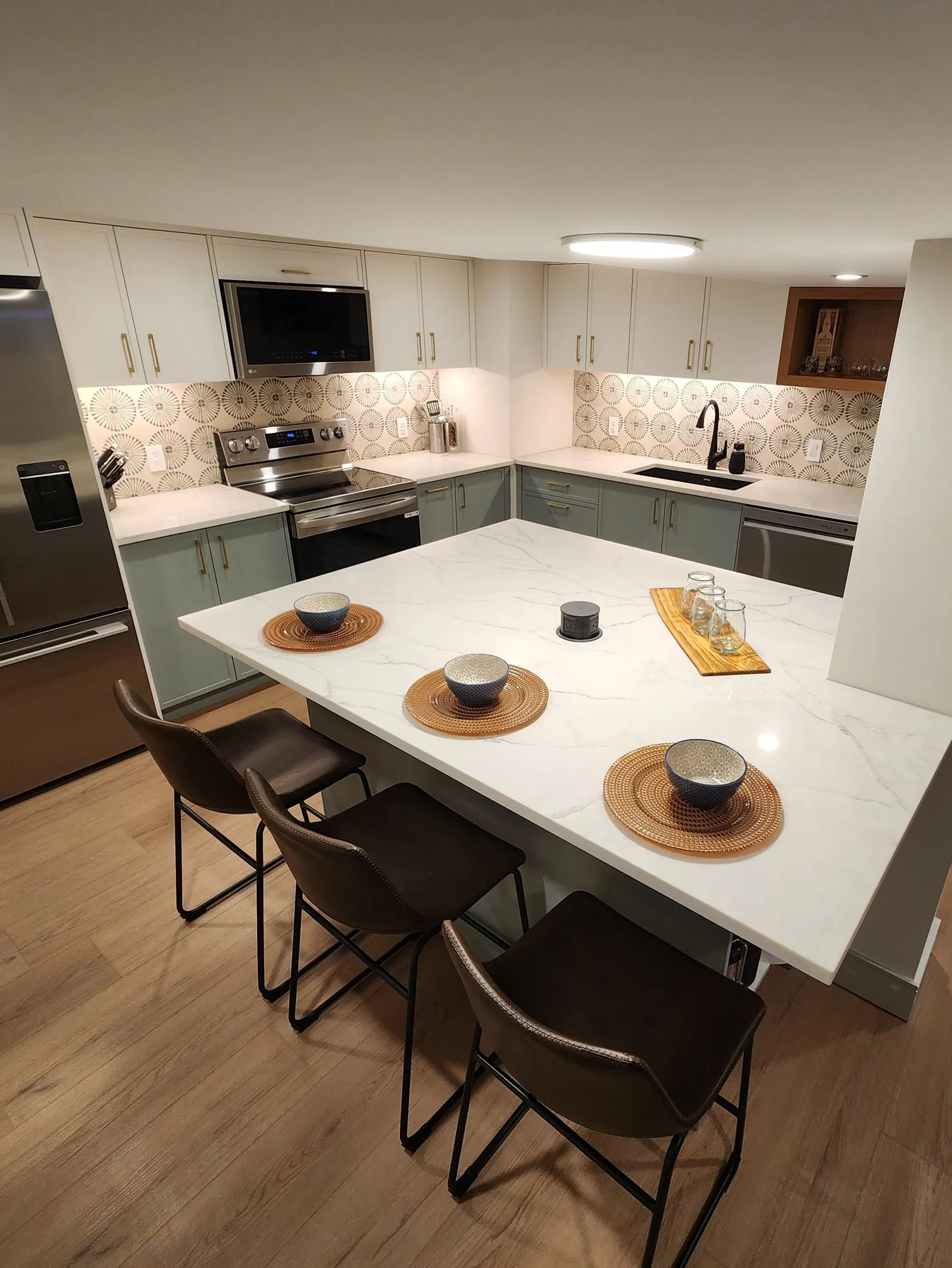 A basement kitchen with sage cabinets, a white island set for dinner and pendant lights